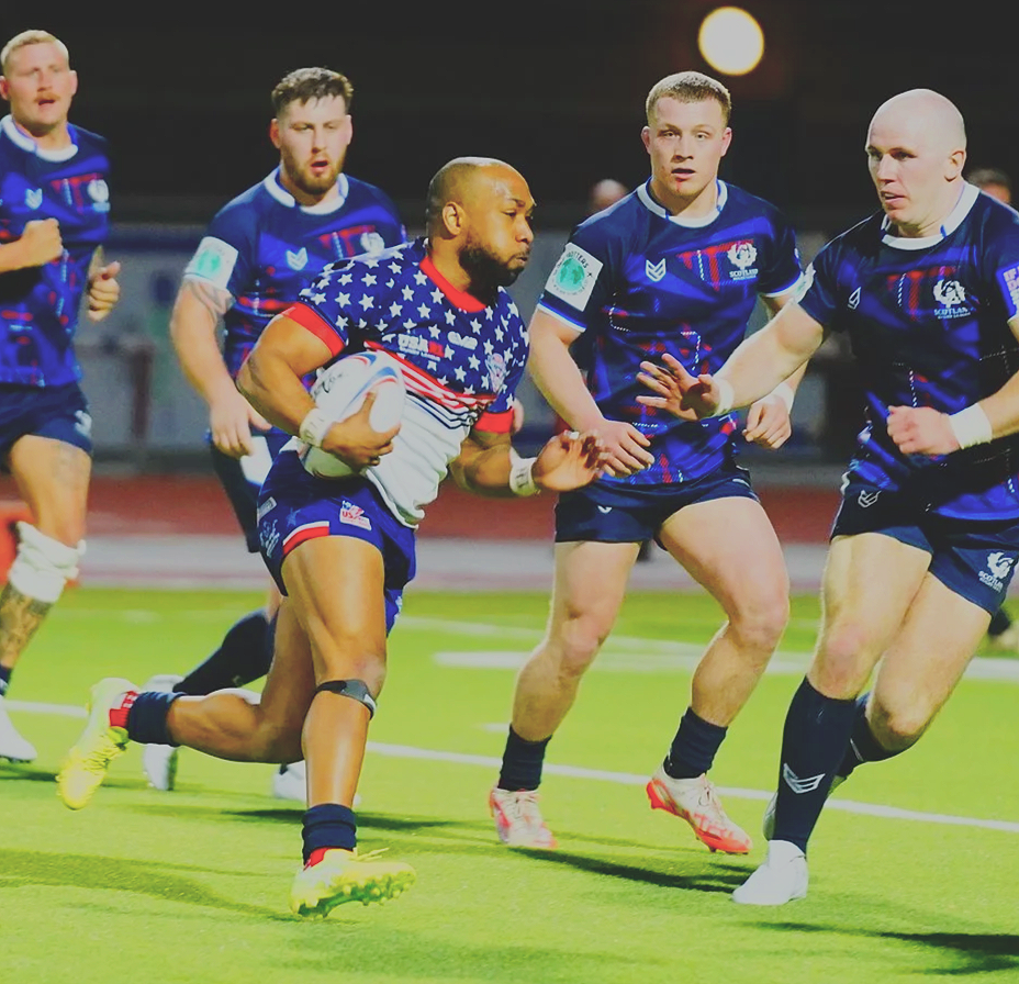 Rugby players in blue jerseys chasing a player in star-spangled jersey holding a rugby ball.