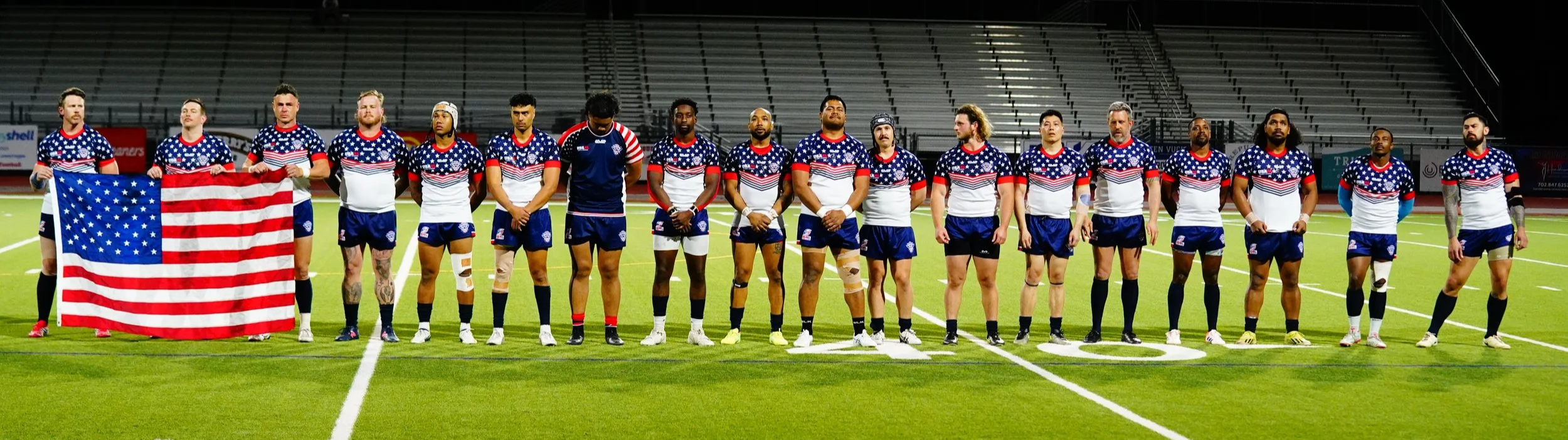 A group of rugby players standing on a field, some holding an American flag, wearing American flag-themed uniforms, in a stadium at night.