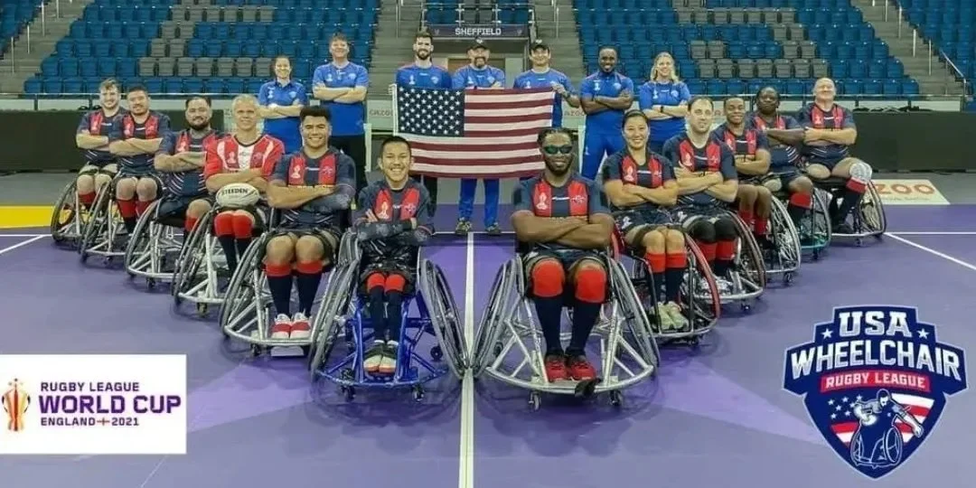 Group of wheelchair rugby players, some in red and black jerseys, some in blue jerseys, posing on a sports court with the USA Wheelchair Rugby League logo and a Rugby League World Cup logo in the foreground, and an American flag in the background.