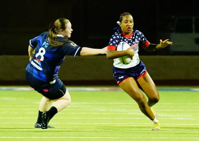 Two female rugby players compete during a game on a field, one of them holding a rugby ball.