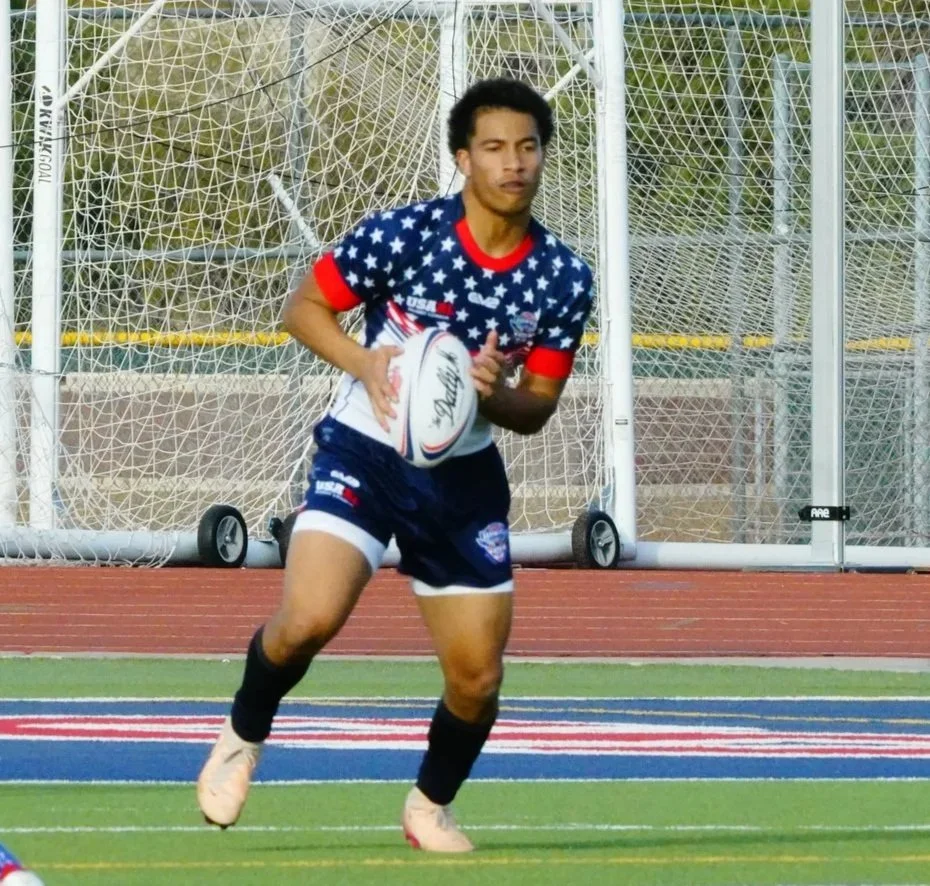 A rugby player in a patriotic USA-themed uniform holding a rugby ball on a field near a goalpost.
