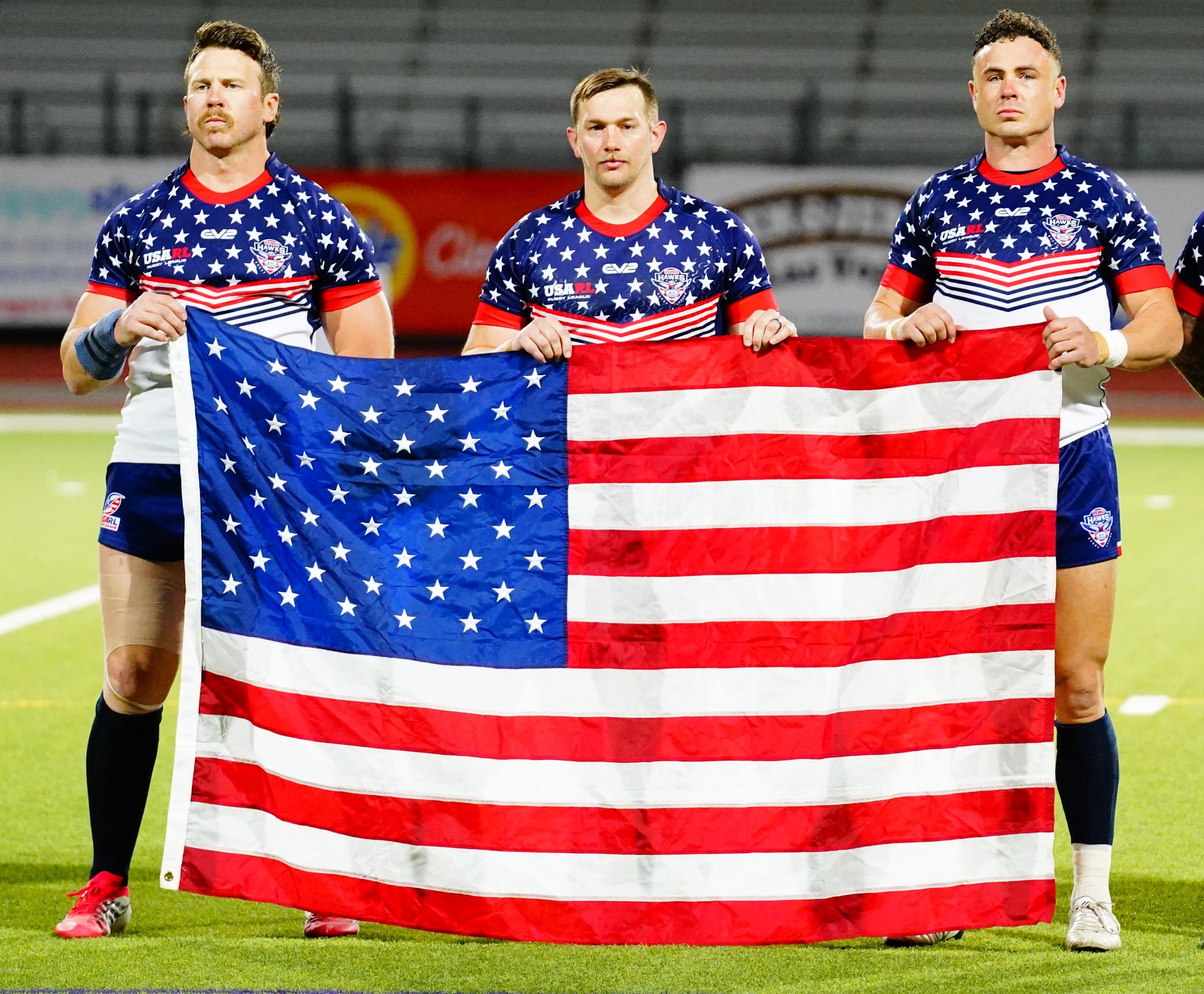 Three male athletes in patriotic sports uniforms holding a large American flag on a sports field, likely before or after a game.