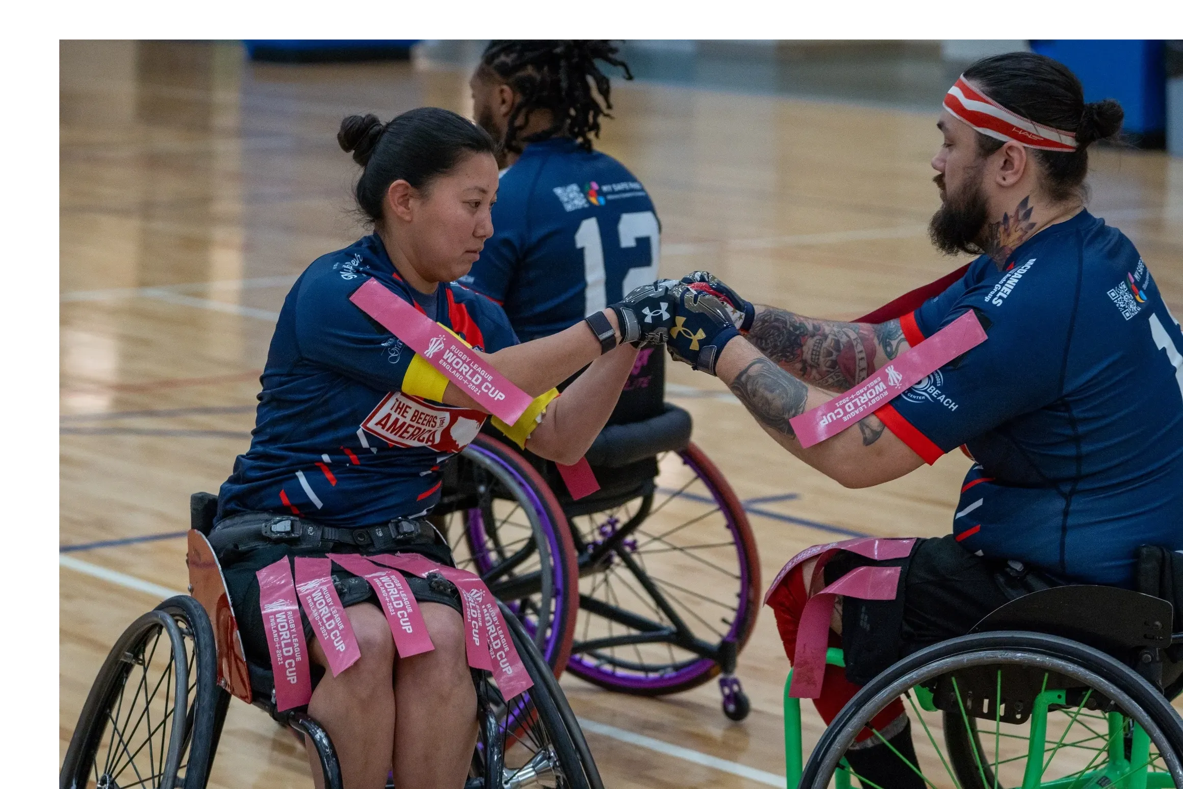 Two wheelchair rugby players, a woman and a man, shaking hands in an indoor sports court. The woman has black hair in a bun, and the man has tattoos, a beard, and a red headband. Both are in blue uniforms with pink wristbands reading "World Cup" and "The Beers Are Legal."