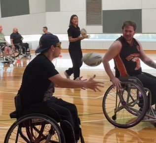 Two women in wheelchairs playing basketball in an indoor gym, with other people in the background.