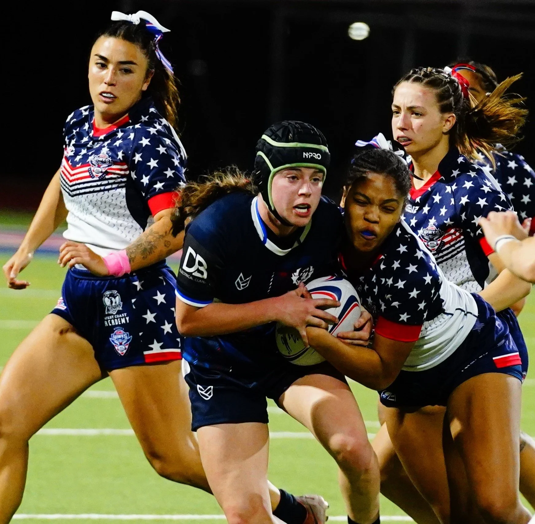 Women playing rugby in a game, wearing patriotic uniforms with stars and stripes, with one player carrying the ball and others attempting to tackle.