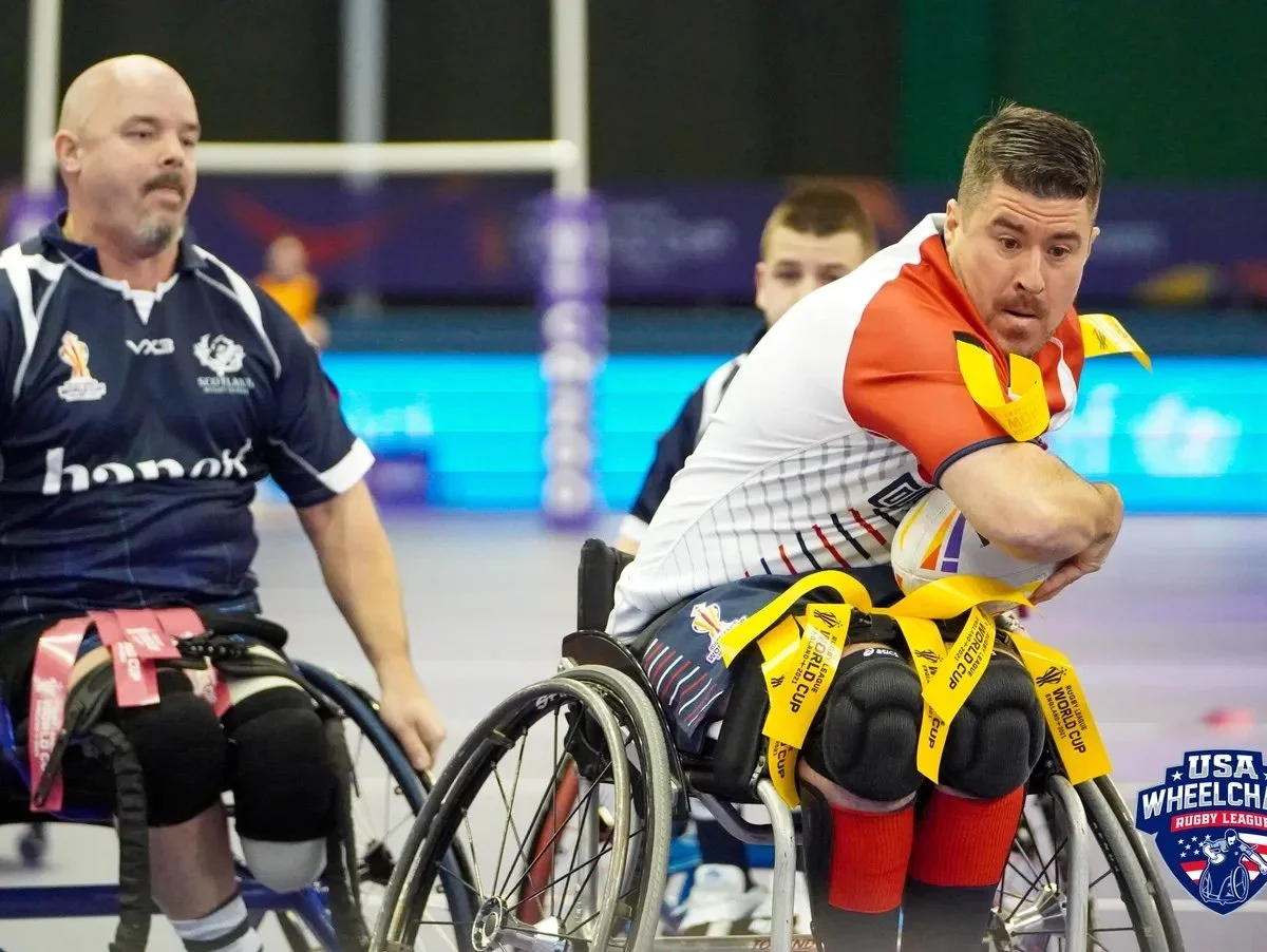Wheelchair rugby players in action during a match, with one player in white and orange aiming to score while others prepare to defend, on an indoor court.