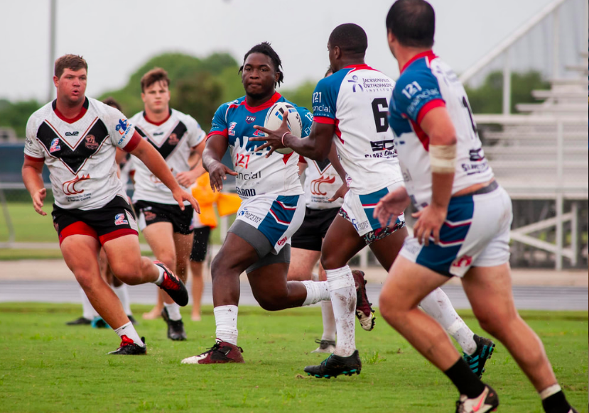 Rugby players in action on the field, with one player running while holding a rugby ball, surrounded by opponents and teammates, some wearing white and others wearing blue and red uniforms.
