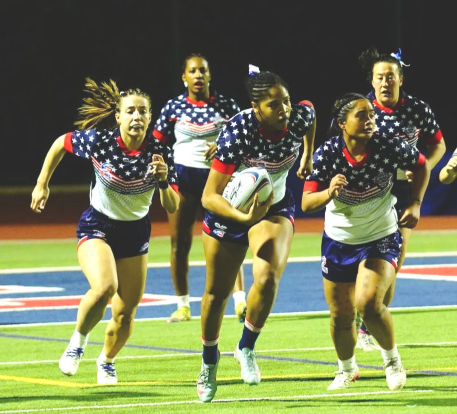Girls playing rugby on a field at night, wearing patriotic red, white, and blue uniforms with stars and stripes.