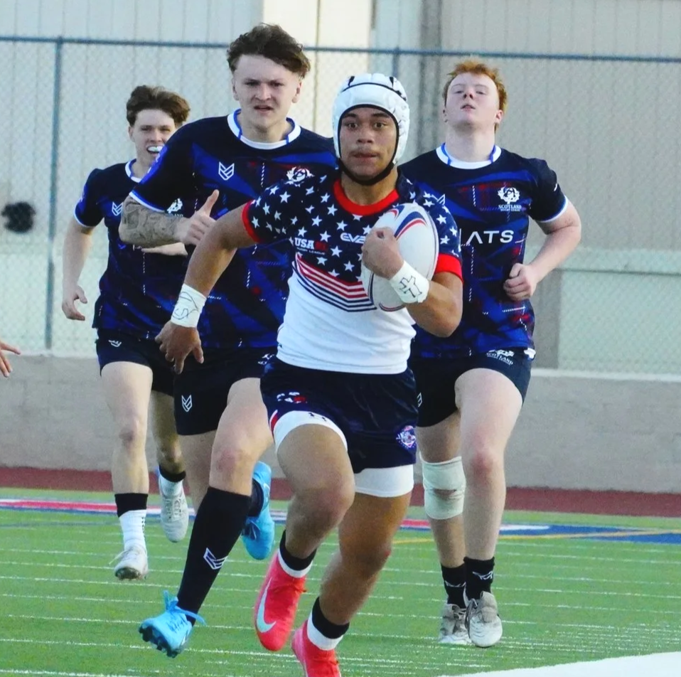 Four rugby players running on the field, with one holding the rugby ball and wearing a helmet, wearing France-themed jersey, and the others wearing team uniforms with Scotland logos, during a game.