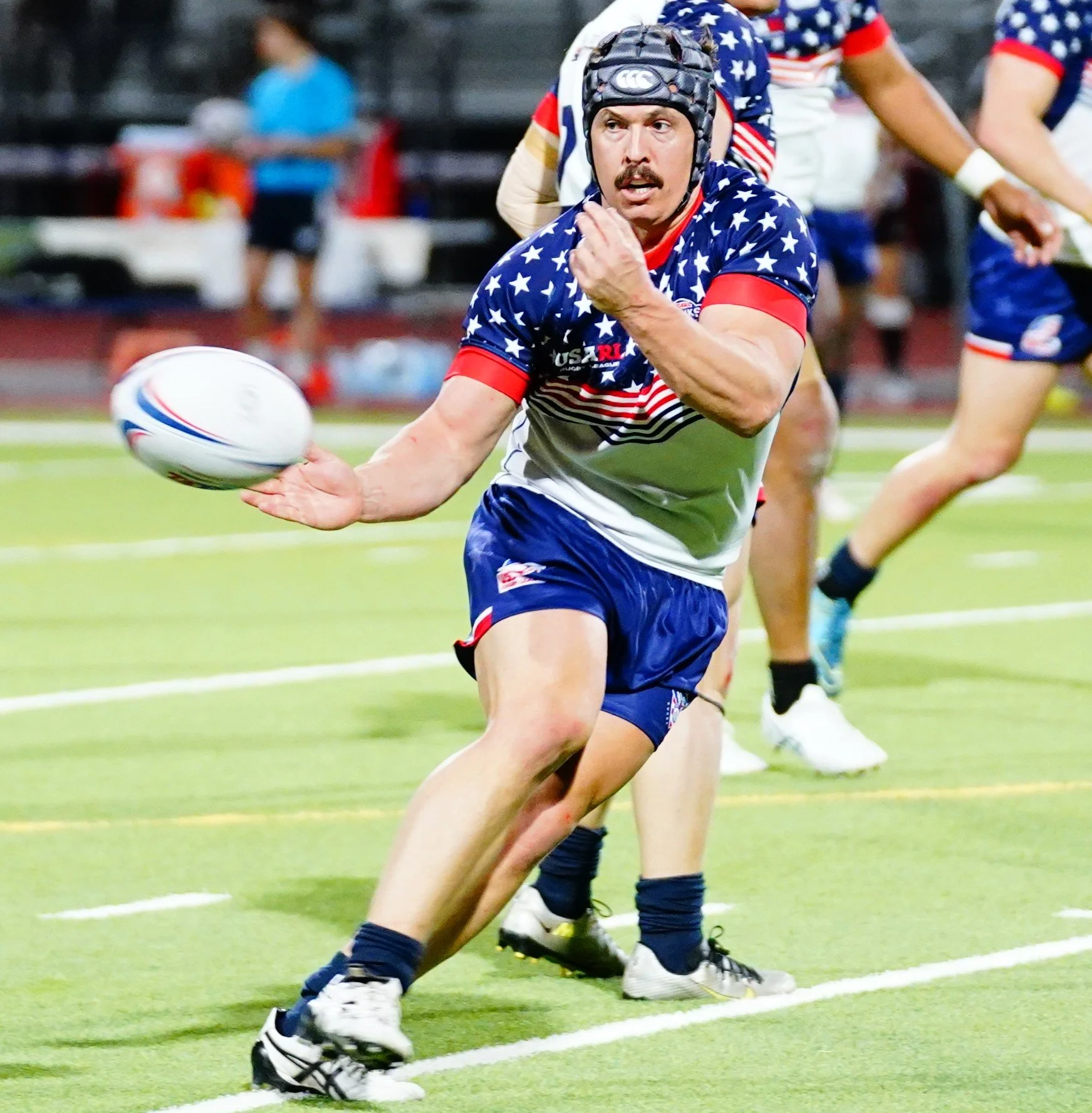 A rugby player in a blue, red, and white jersey with stars, running on a field with a rugby ball in hand, wearing a helmet, and surrounded by teammates.