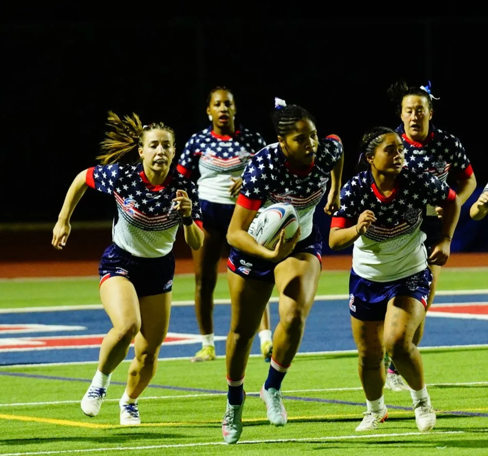 Group of female rugby players in patriotic uniforms running on a field at night, with one holding a rugby ball.