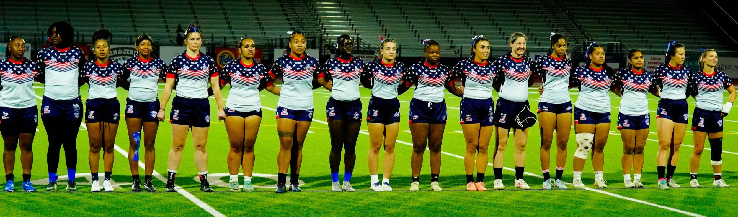 A group of female athletes standing in a line on a football field, wearing matching uniforms with American flag-themed shirts and shorts, some with protective gear, during a sports event at night.