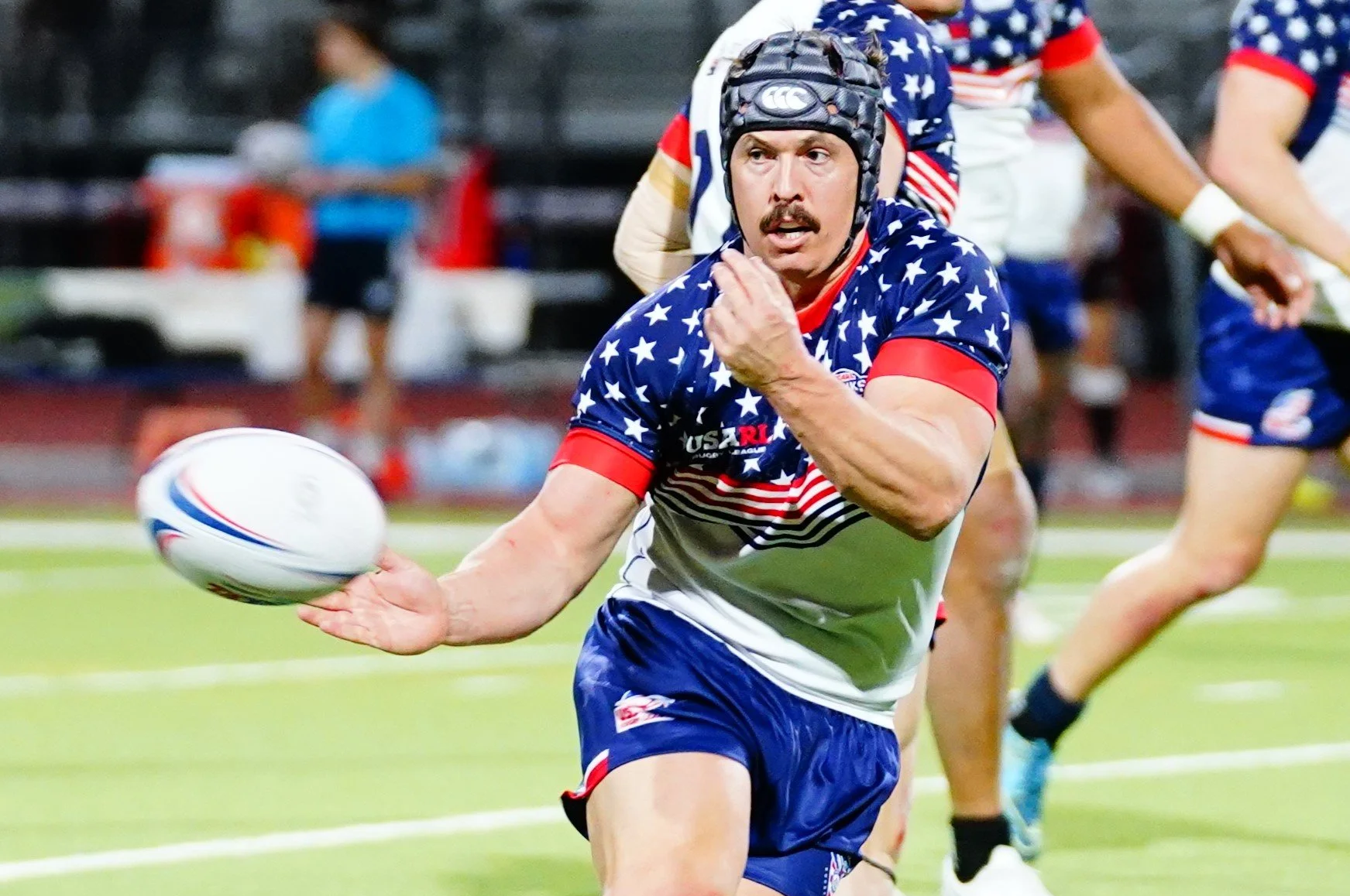 Man playing rugby in American flag-themed jersey and helmet, holding a rugby ball during a game.