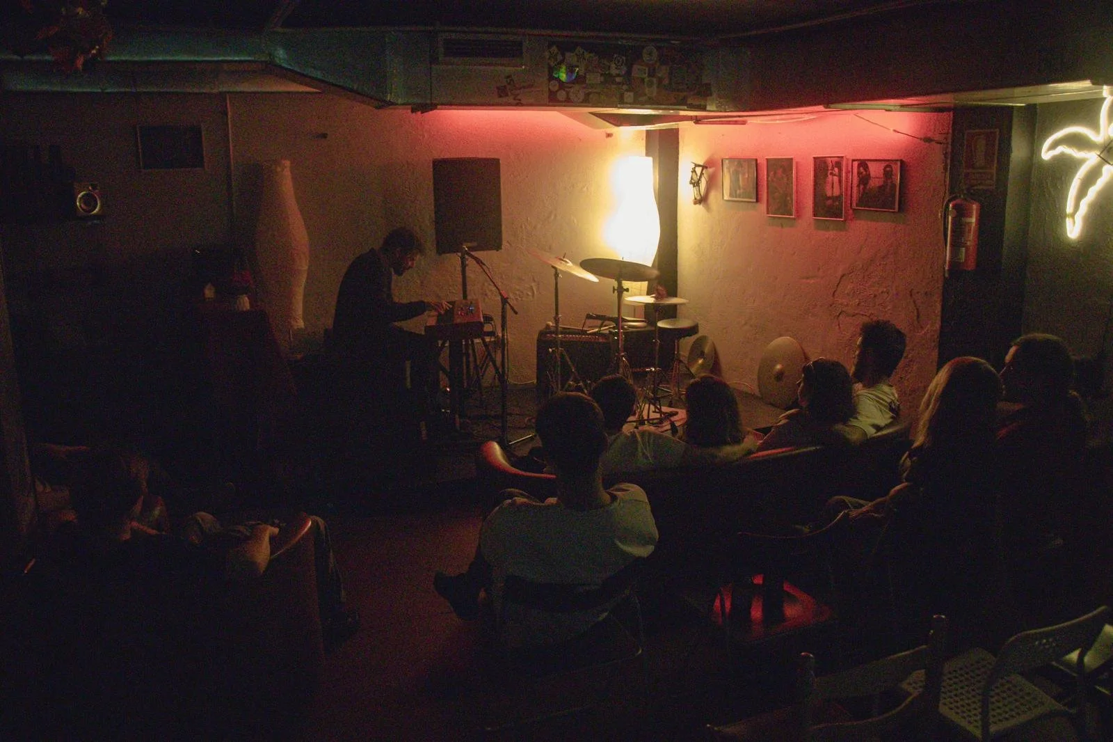 A dimly lit underground music venue with a small stage and an audience seated on chairs and sofas, watching a performer playing a keyboard. The stage has a microphone, drums, and music equipment, with artwork on the wall and neon lights.