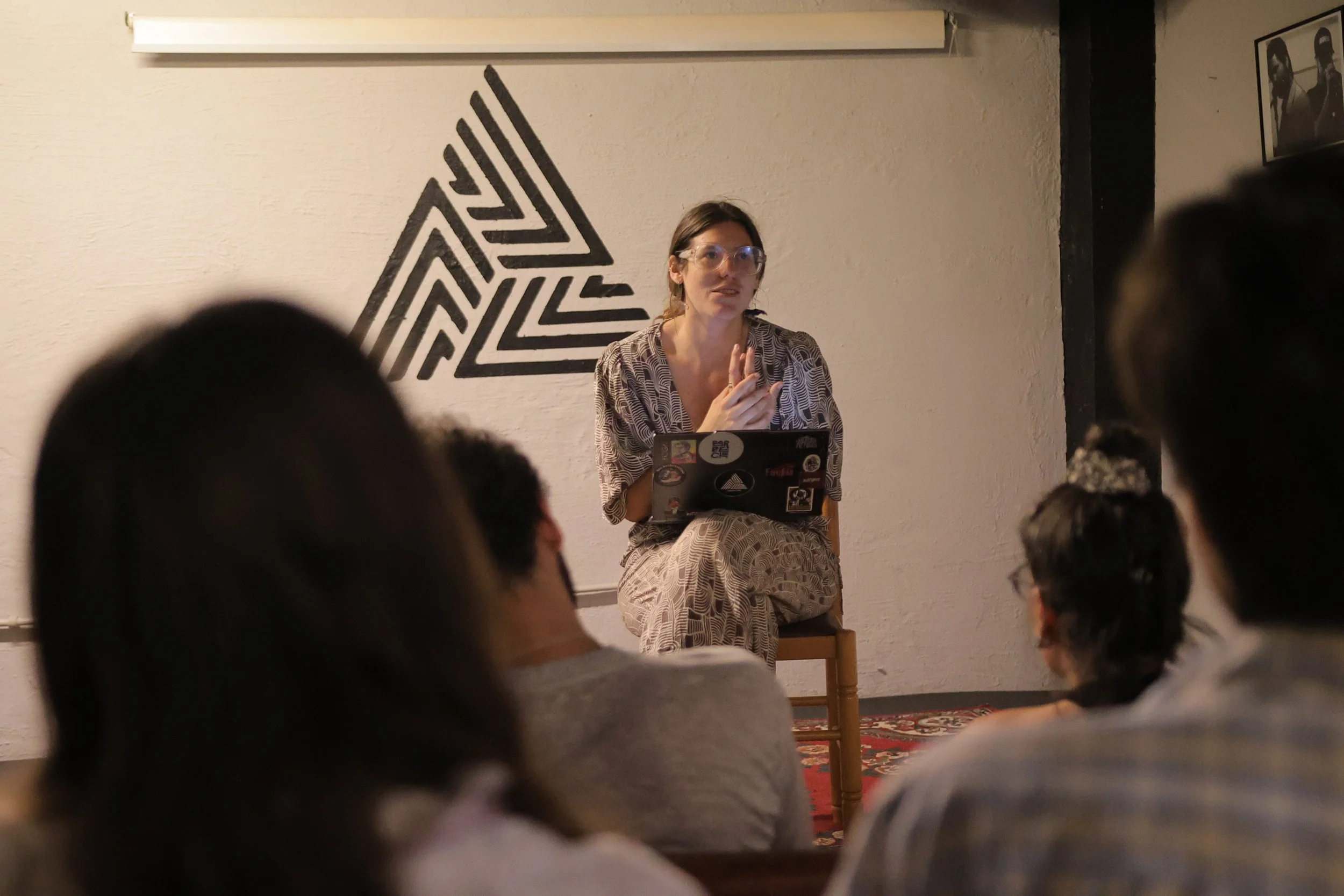 A woman sitting on a chair with a laptop on her lap, speaking to an audience in a small room, with a large geometric black and white wall art behind her. Several people are listening, with their backs to the camera.