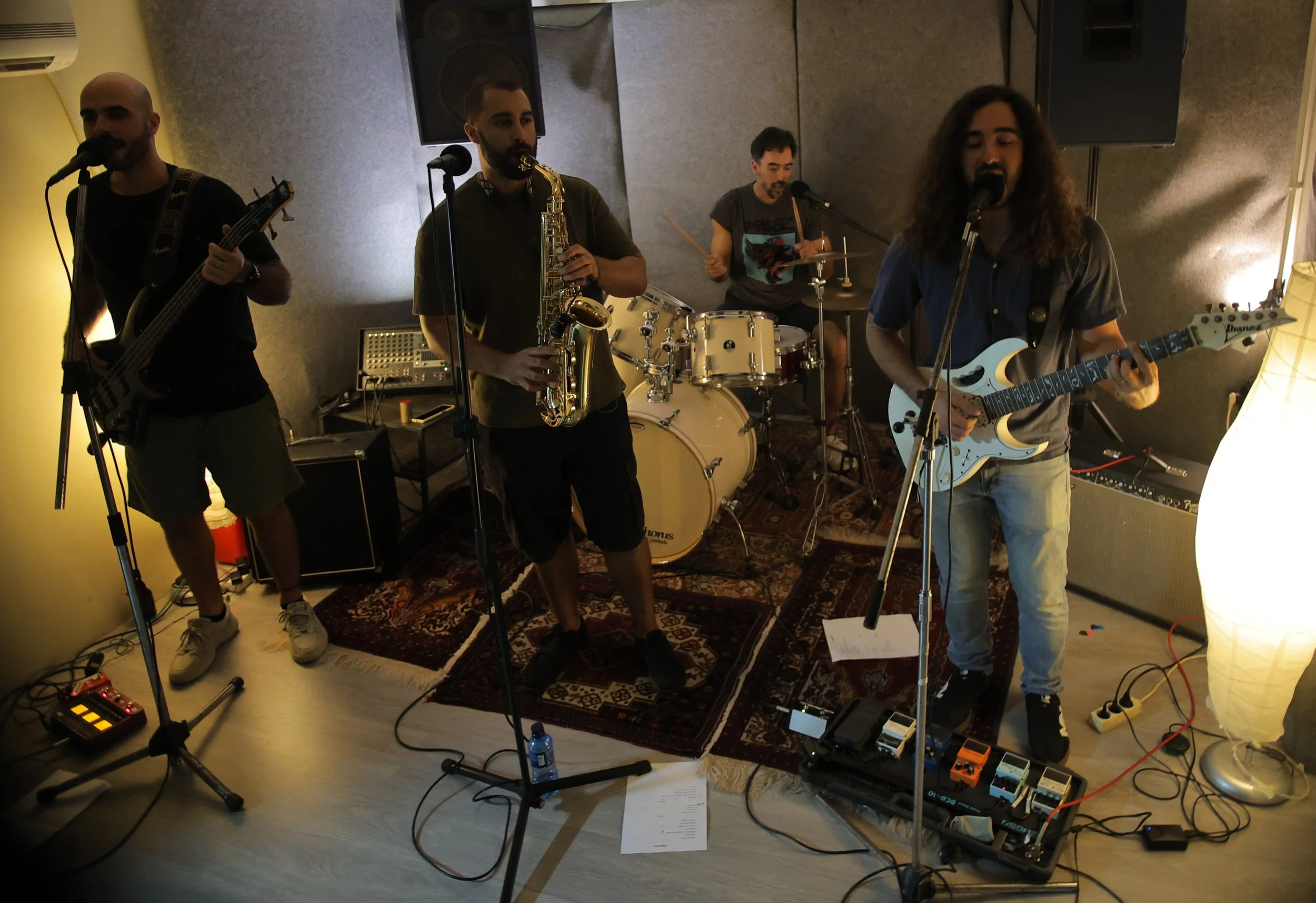 A band of four musicians performing indoors. The band includes a guitarist, a bassist, a saxophonist, and a drummer. They are using microphones and various musical equipment, with a patterned rug on the floor and a lamp nearby.