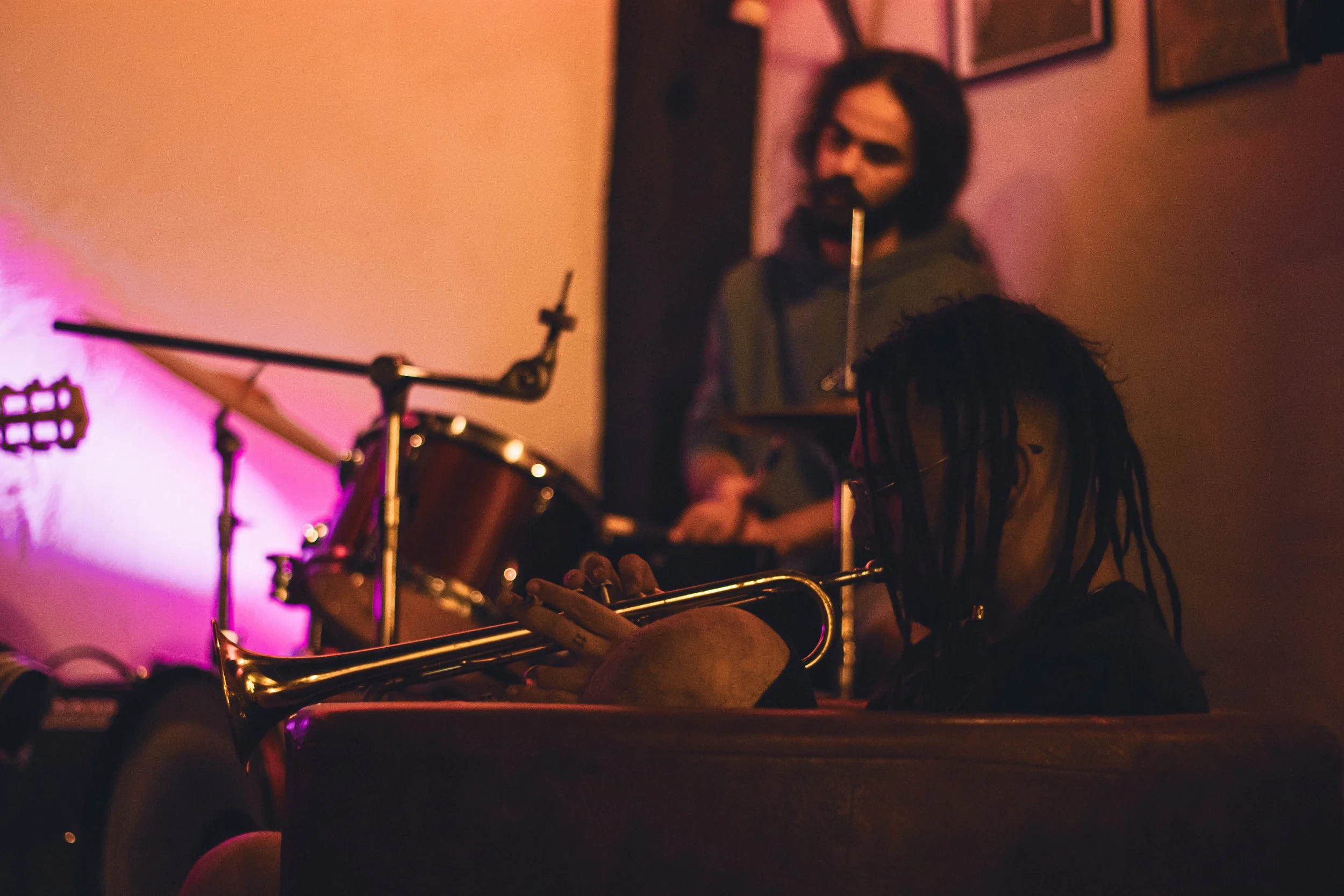 A person with dreadlocks and glasses sitting with a trumpet, in a dimly lit room, with a drummer in the background playing drums.