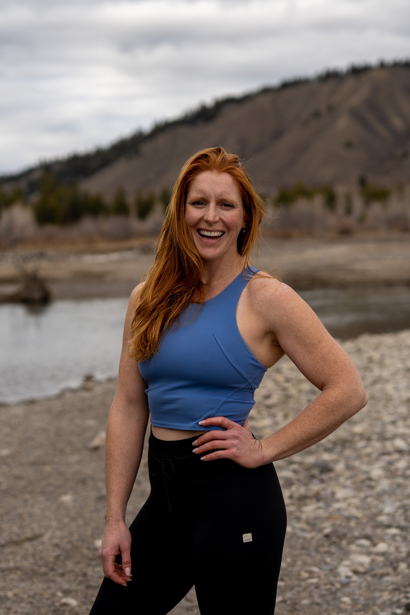 A smiling woman with long red hair in athletic clothing, standing outdoors near a river and mountains.