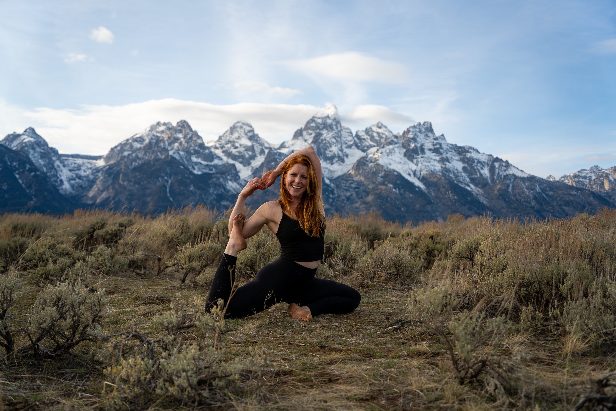 A woman practicing yoga outdoors in a field with mountains in the background.
