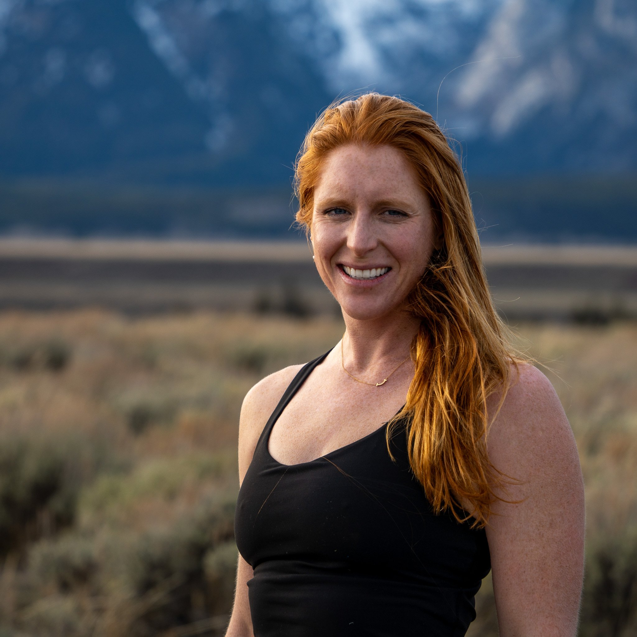 A woman with red hair, wearing a black tank top, smiling outdoors with a mountain range and a cloudy sky in the background.