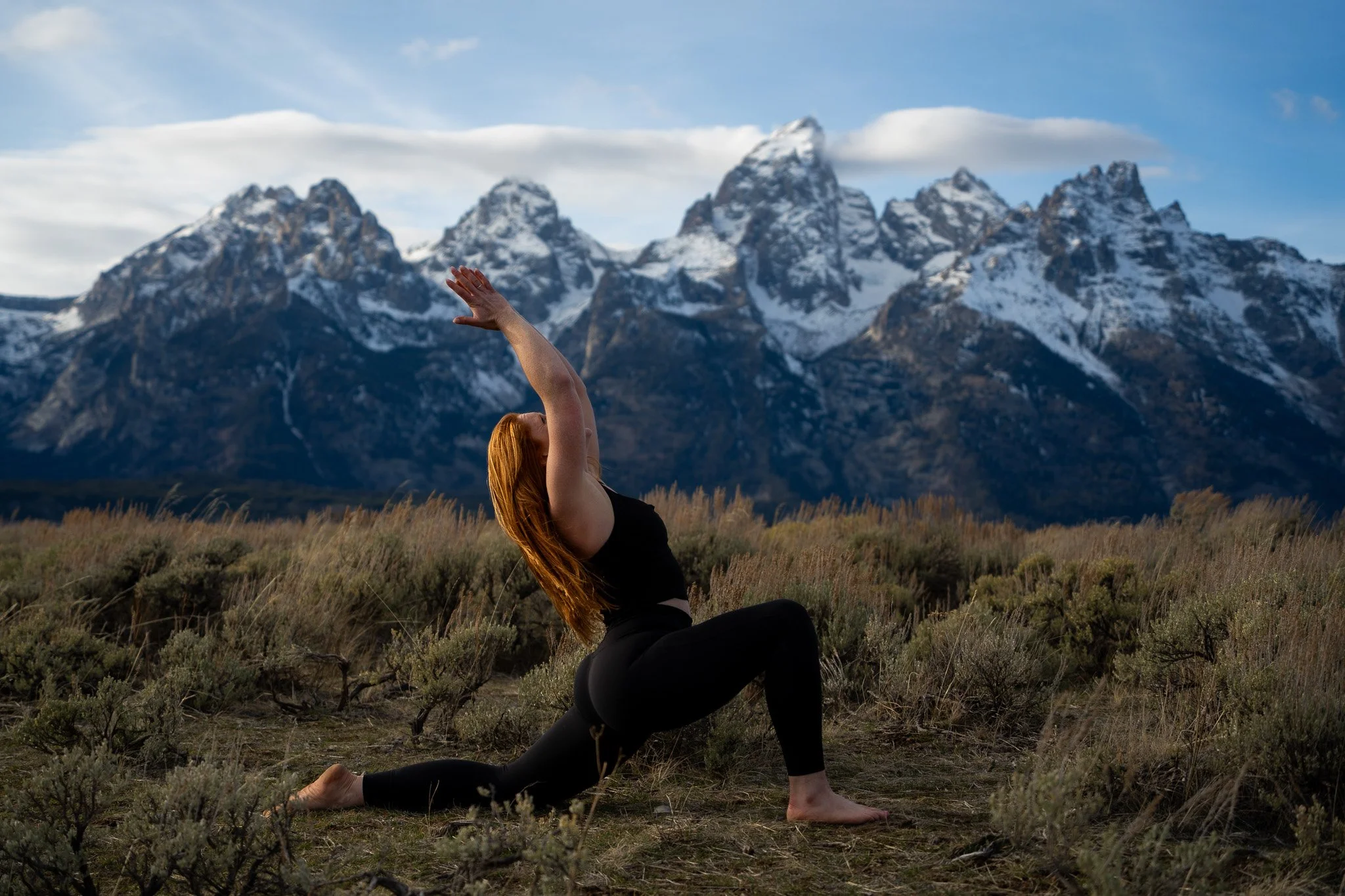 A woman practicing yoga outdoors in a natural field with mountains and snow in the background.