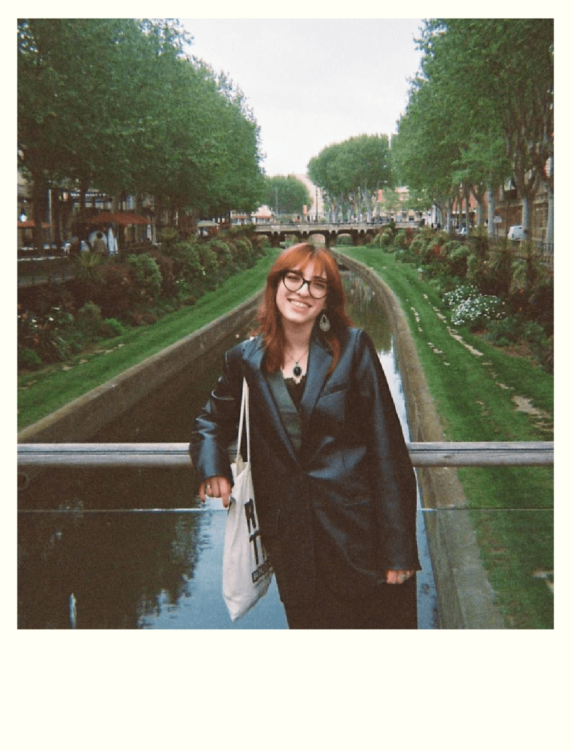 A woman with red hair and glasses smiling, standing near a railing over a small water channel in a park or city area with trees, bushes, and a bridge in the background.