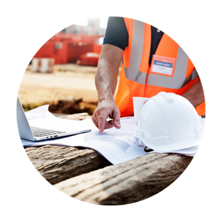 Construction worker in orange safety vest and black shirt pointing at blueprints on a wooden surface with a white safety helmet nearby.