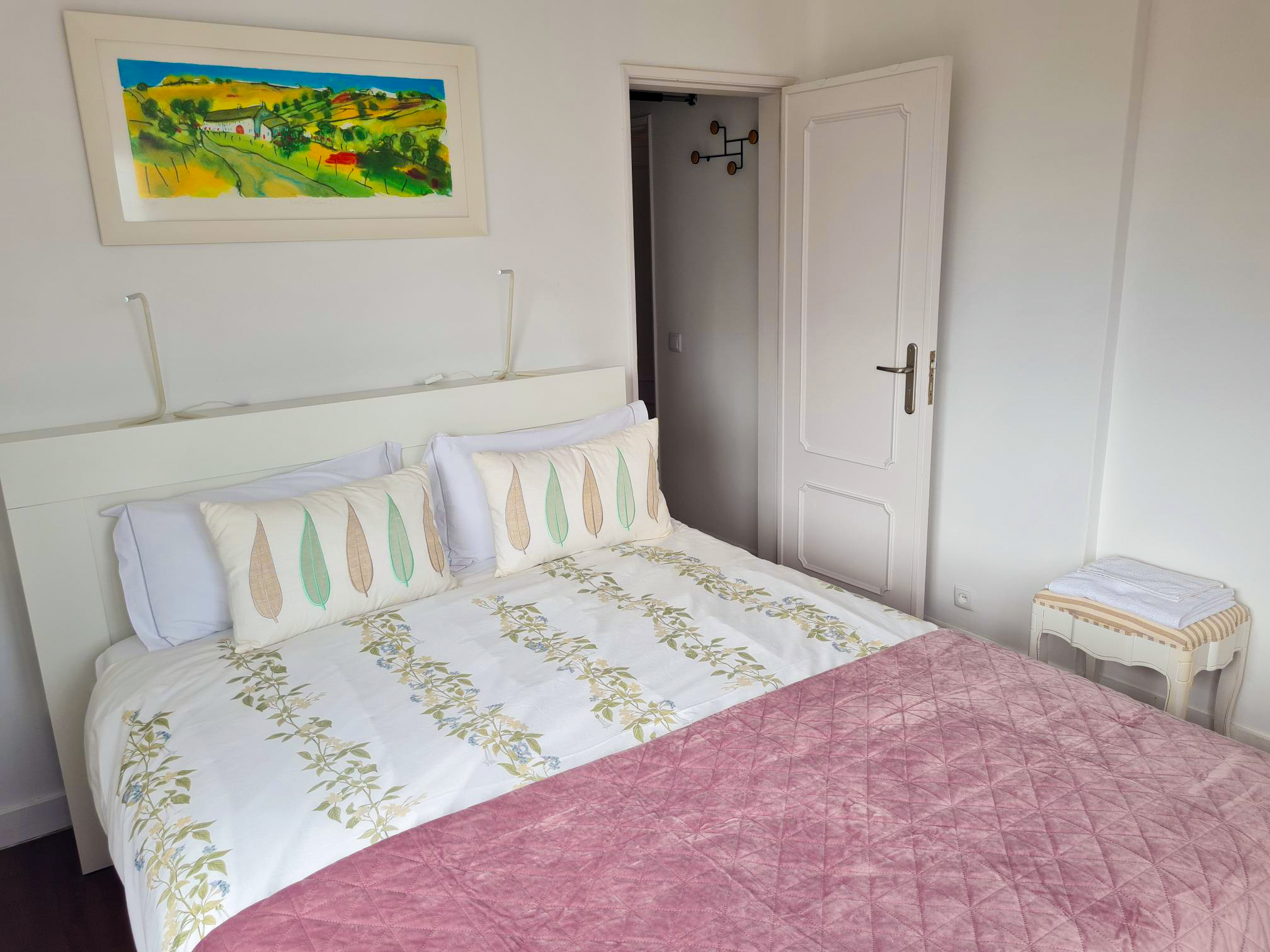A neatly made bed with white pillows and floral patterned pillowcases, a floral bedspread, and a pink quilt. Above the bed, a colorful landscape painting hangs on the white wall. To the right, a small table with folded towels is beside a closed white door with trim and a metallic handle. The room is brightly lit with minimalist decor.