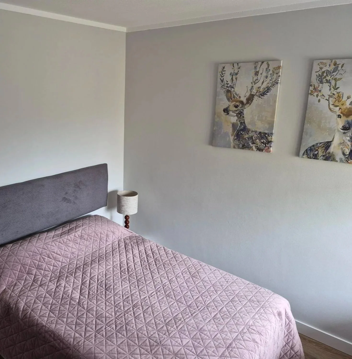 A bedroom with a bed covered in a dark green textured blanket, three white pillows with embroidered edges, a grey upholstered headboard, a small side table with a red and beige lamp, and a black and white geometric rug on wood flooring.