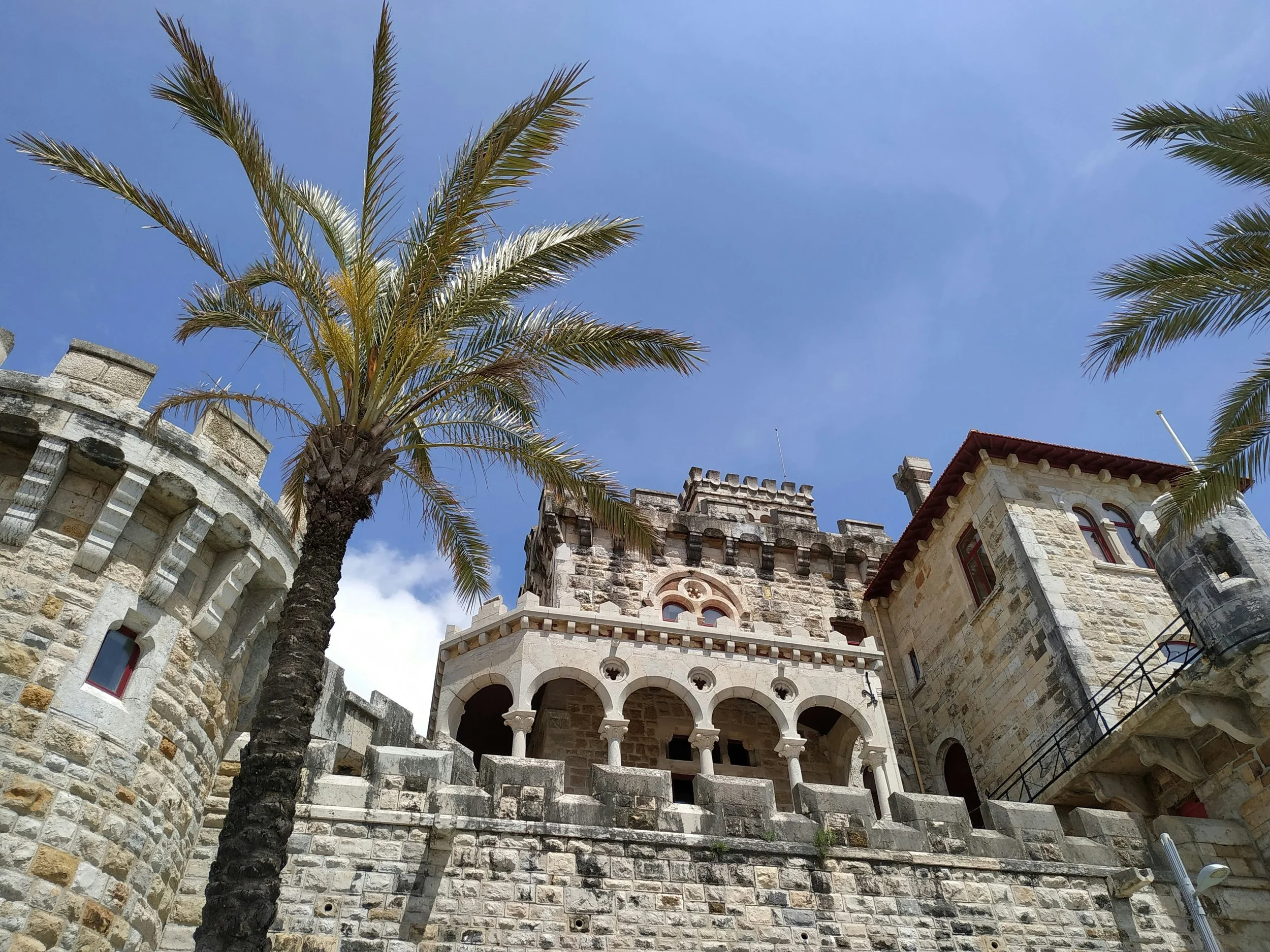 Stone castle-like building with arches and towers under a blue sky, flanked by palm trees.