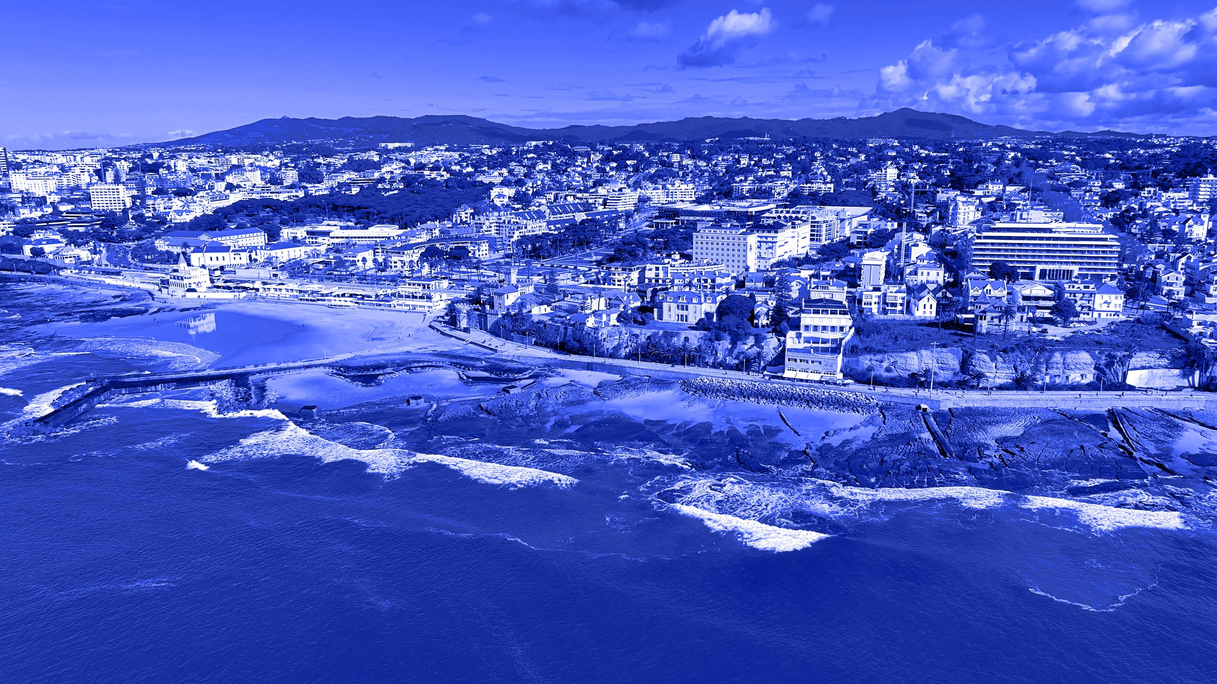 A panoramic aerial view of a coastal city with buildings, a beach, and rolling hills in the background, under a partly cloudy sky.