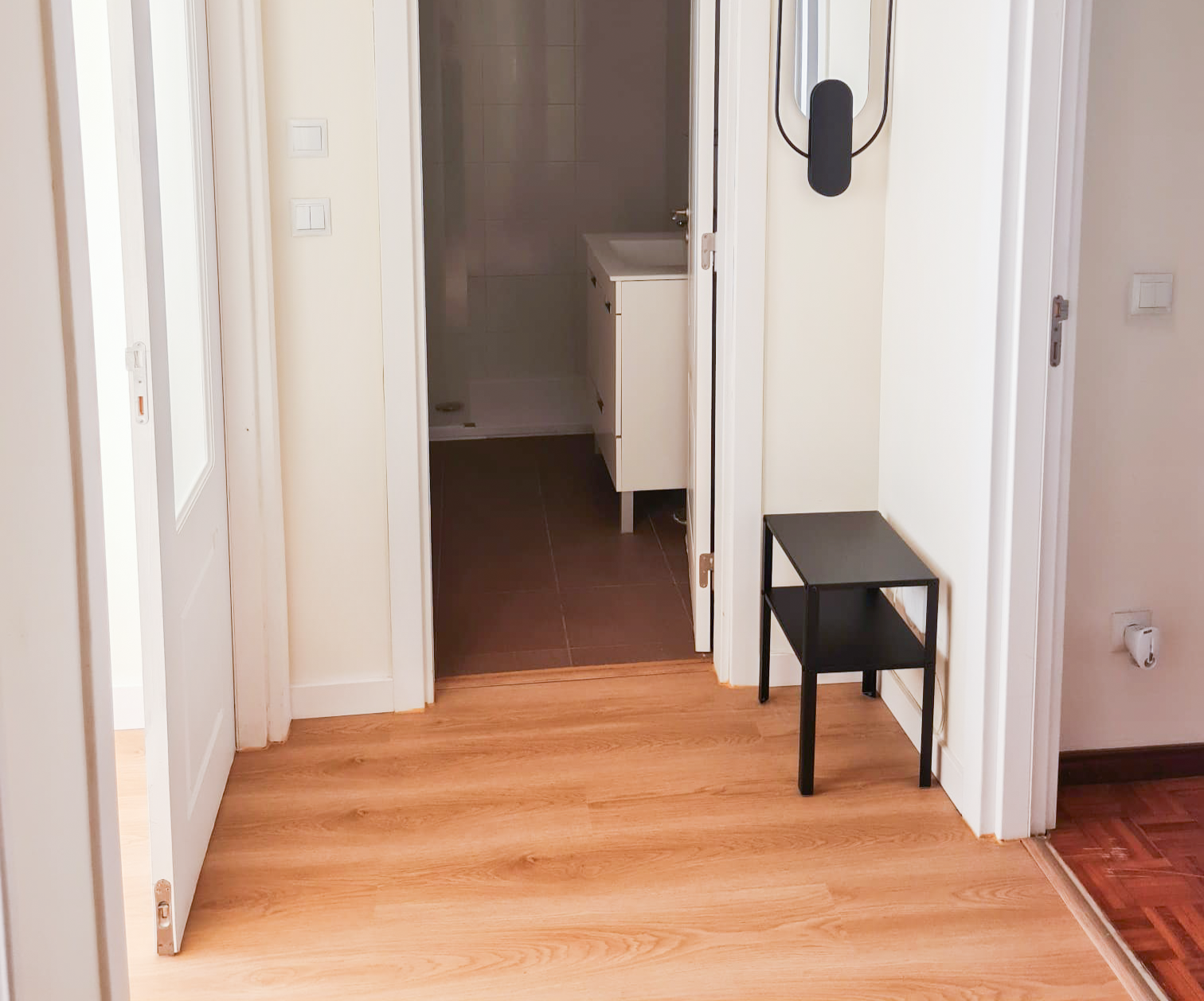 View of a hallway with light wood flooring, leading to a bathroom with dark tile flooring. There is a small black side table and a wall-mounted light fixture.