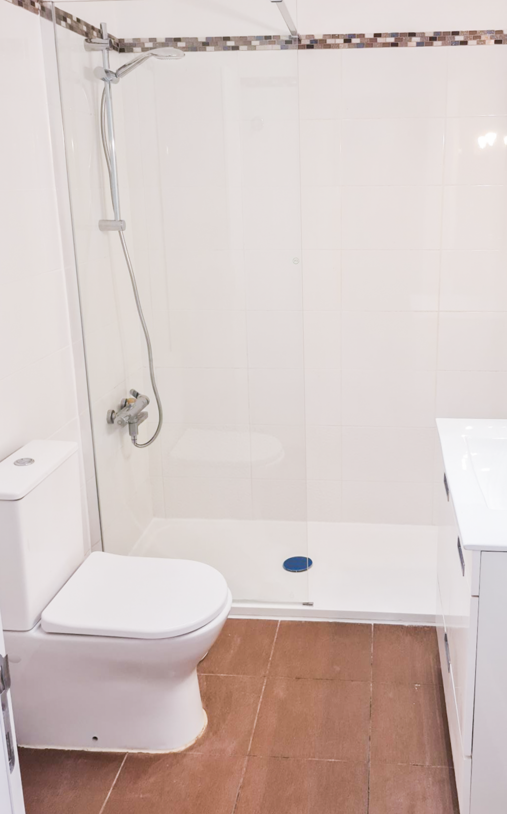 Small bathroom with a toilet, a shower area with a glass door, and a white sink cabinet, brown tiled floor, white tiled walls.
