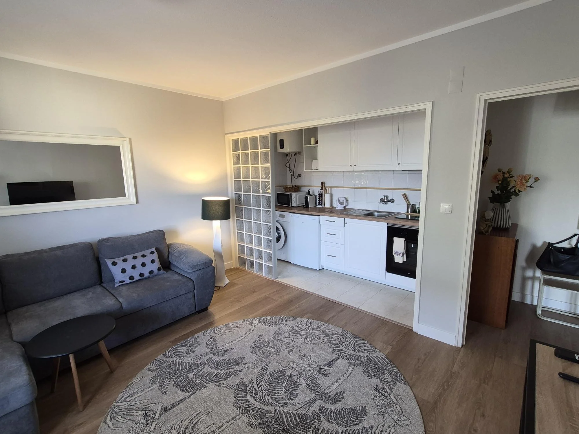 Small kitchen with white cabinets, wooden countertop, and oven, sink, microwave, and washing machine. Separate area with glass block wall, potted plant, and a sofa in the foreground.