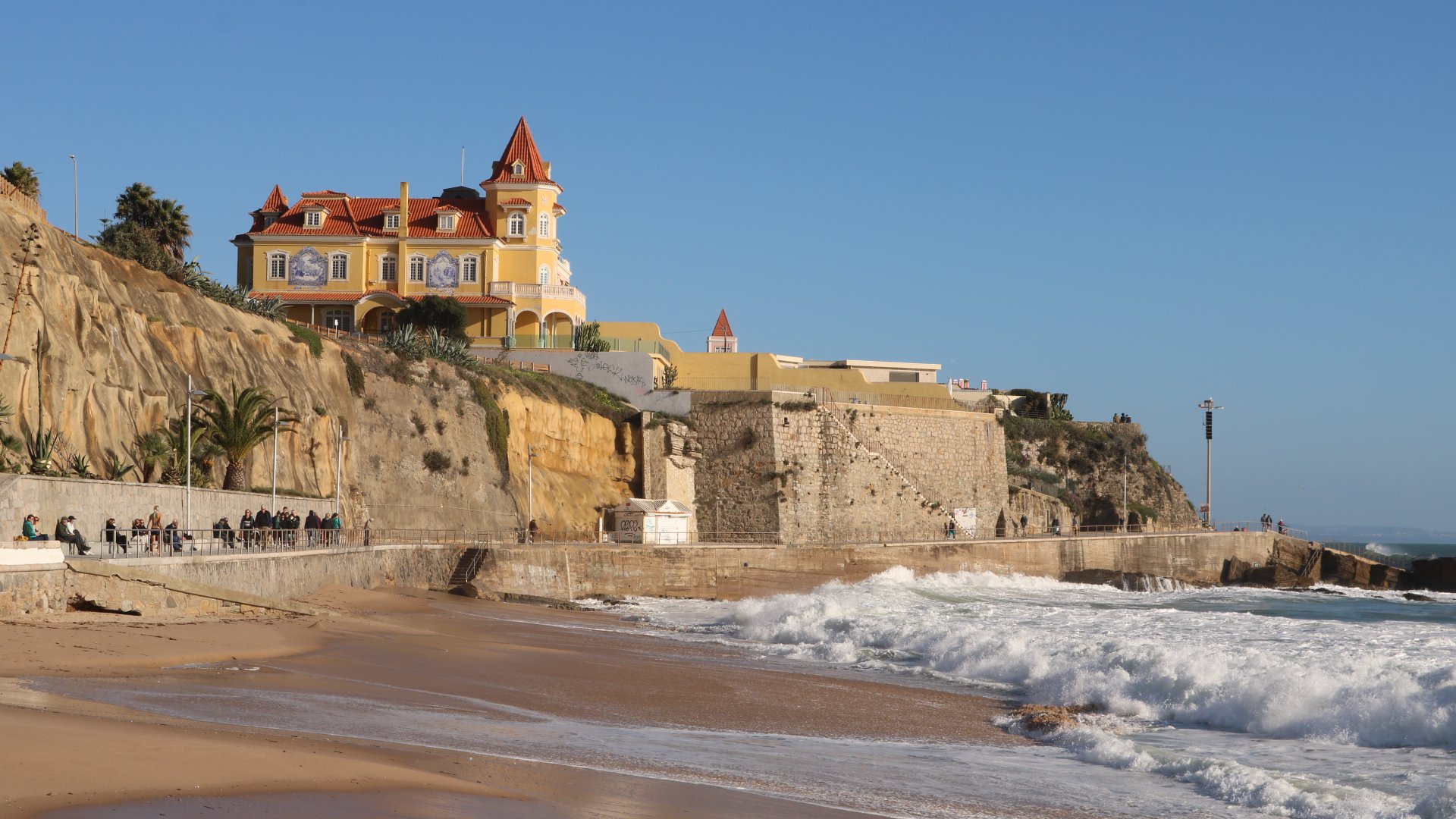 Beachside promenade with a yellow and orange castle-like building on a rocky cliff, ocean waves crashing on the shore, and a few people sitting on benches and walking along the coast on a clear day.