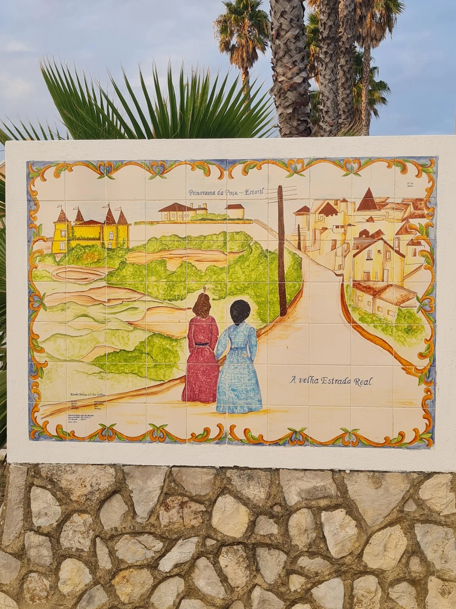 Decorative ceramic tile mural depicting a scenic view of a historical town, with two women in period clothing walking along a path, surrounded by greenery and old buildings, framed with colorful ornamental borders.