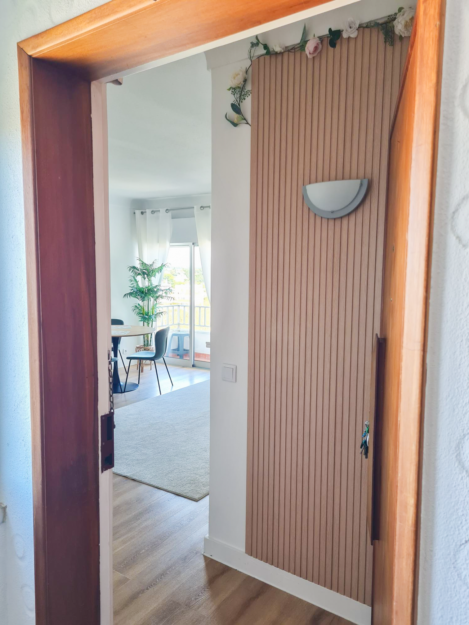 View through a doorway into a bright living space with a dining table, chairs, a large window with curtains, a potted plant, and outdoor balcony railing.