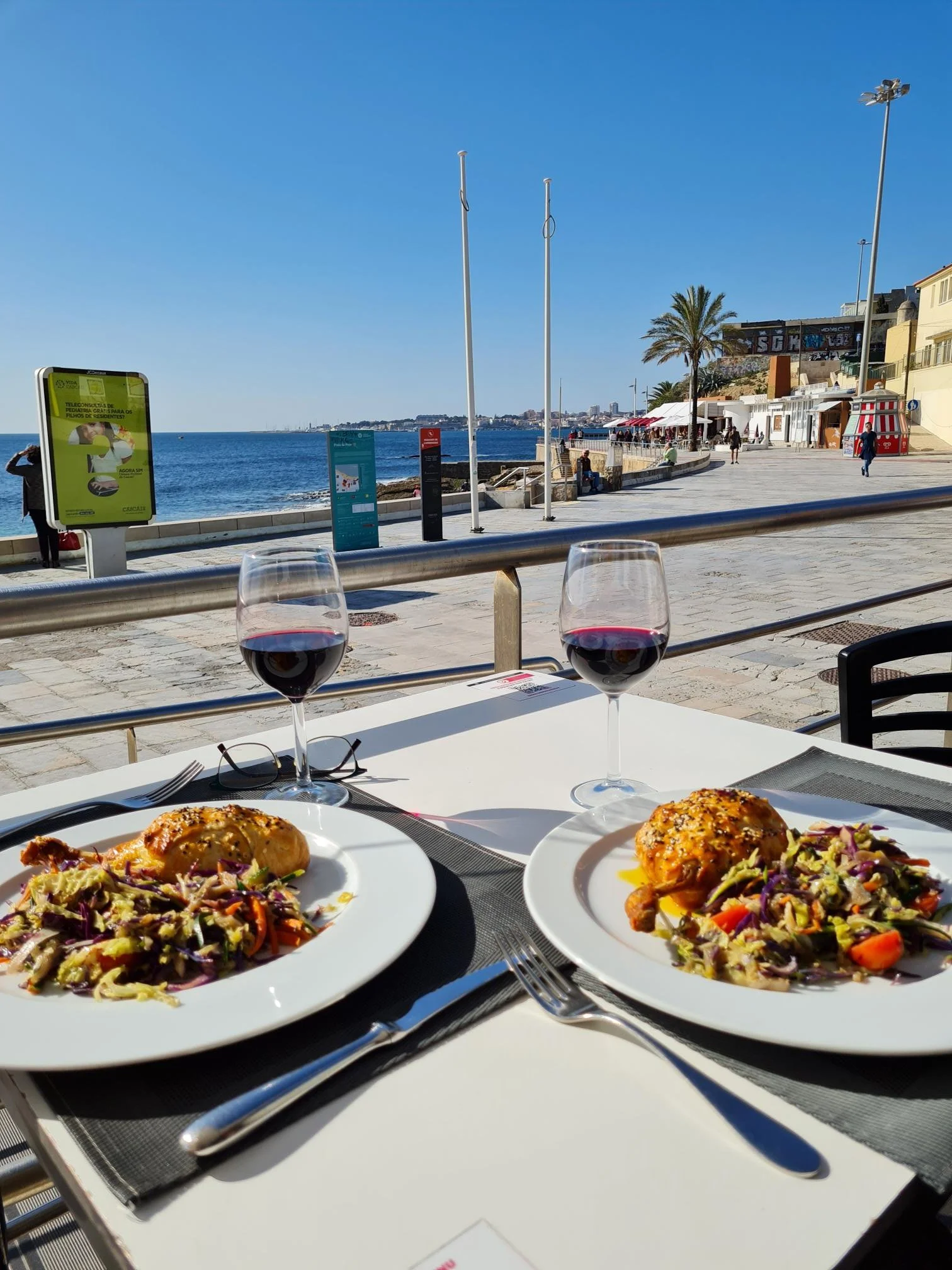 Two plates of food with roasted chicken and vegetable salad, two glasses of red wine, and a pair of glasses on a white table at an outdoor restaurant by the ocean, with a walkway, palm trees, and buildings in the background on a sunny day.