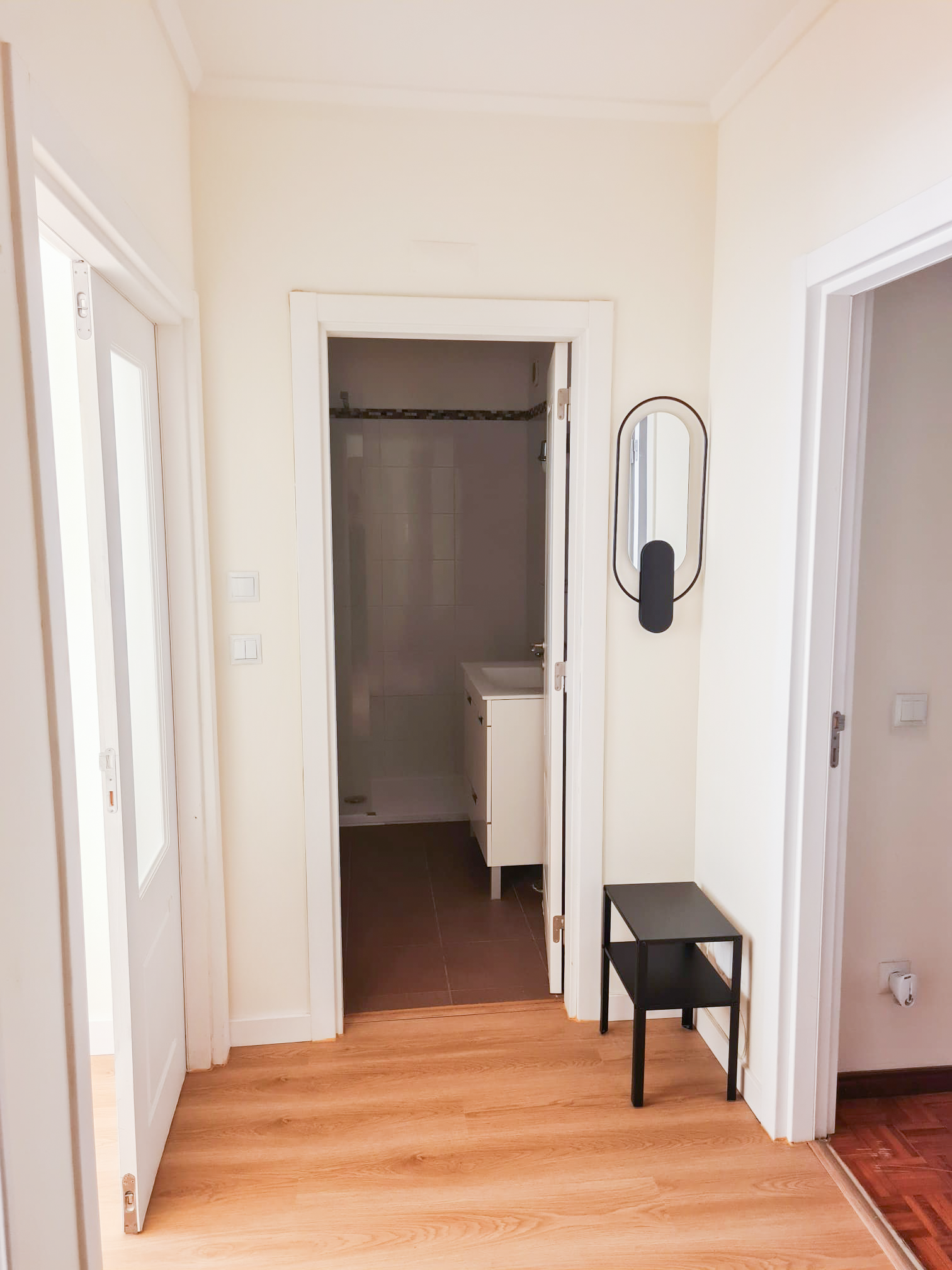 View of a hallway with light wood flooring, leading to a bathroom with dark tile flooring. There is a small black side table and a wall-mounted light fixture.