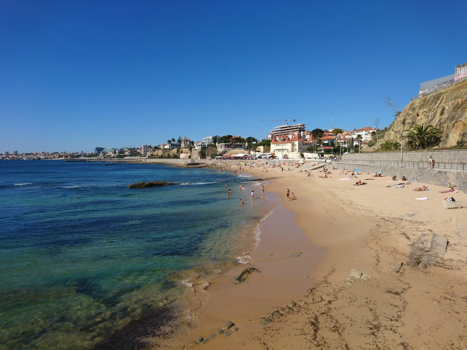 A sunny beach with turquoise water, swimmers, and people sunbathing on the sand. There are houses and buildings on a hill in the background under a clear blue sky.