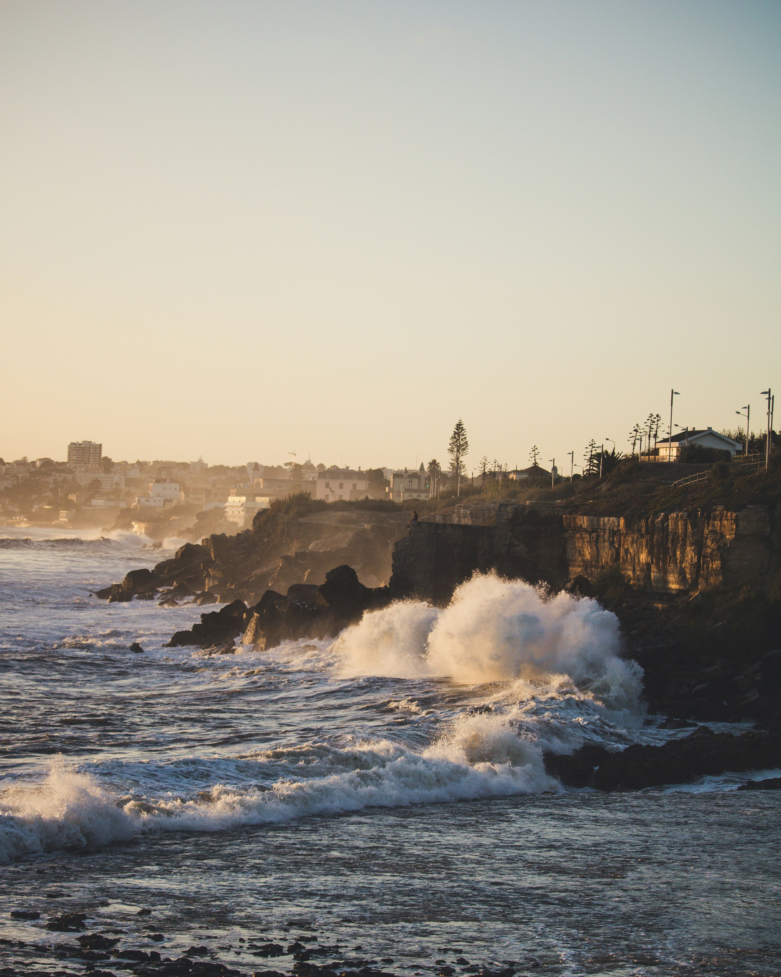 Sunset over rocky coastline with waves crashing, buildings and trees in the distance.