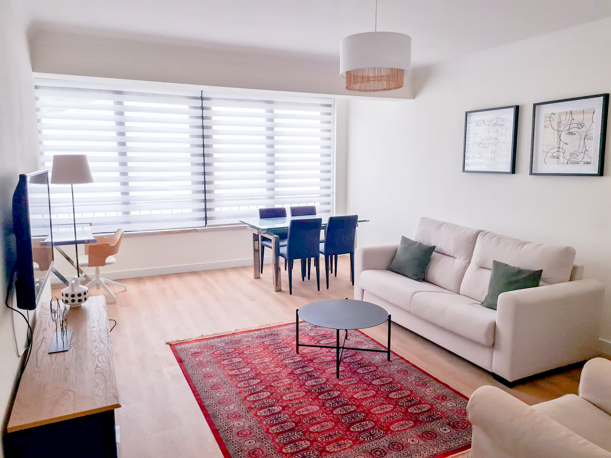 Bright living room with large window blinds, a white sofa with green pillows, a red patterned rug, a round black coffee table, wall art, a dining table with dark chairs, a desk with a white chair, a TV, and a modern ceiling light.