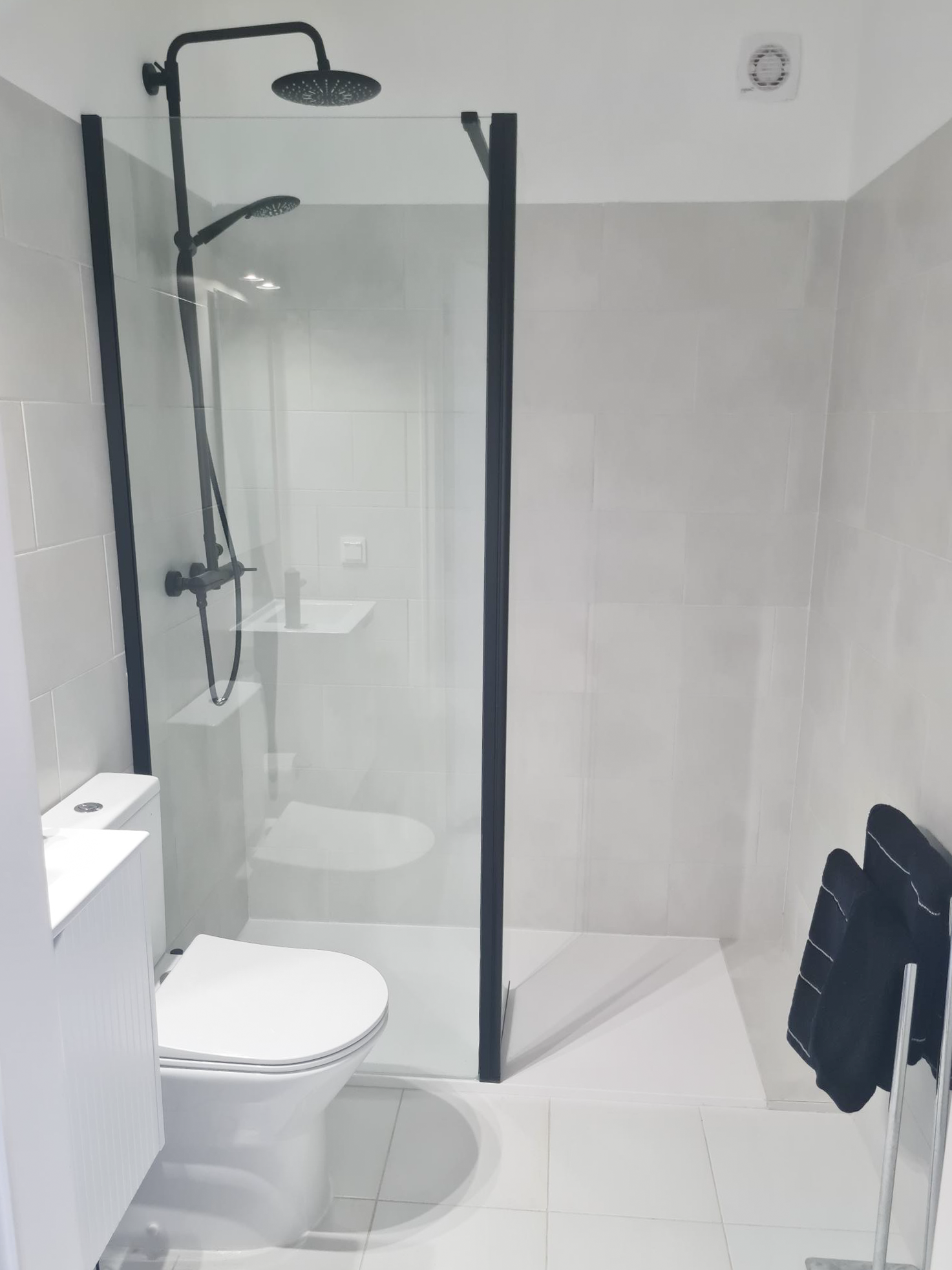 Modern bathroom with walk-in shower enclosed by glass, black shower fixtures, a small white sink, and a black towel hanging on a rack.