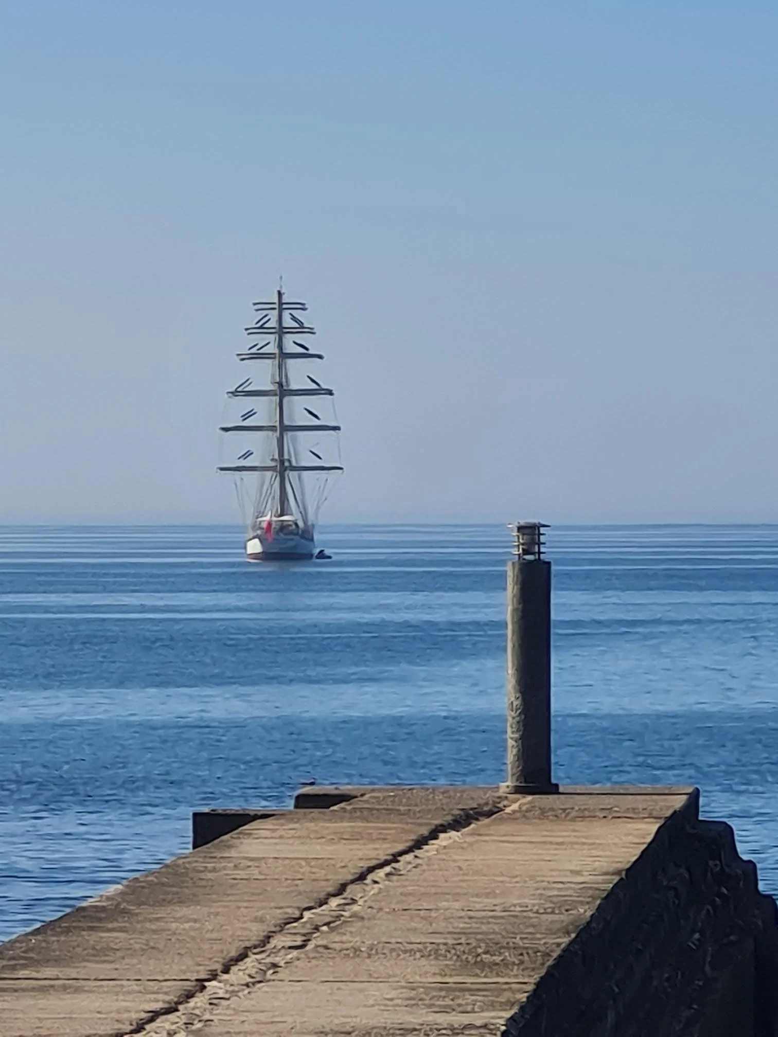 A large sailing ship with multiple masts is sailing on calm blue water, approaching a concrete pier or breakwater that extends into the water. The sky is clear with no visible clouds.