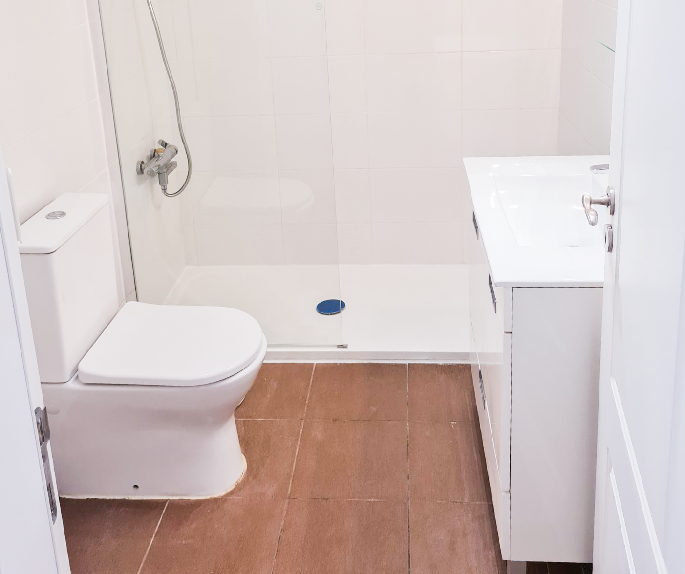 Small bathroom with a toilet, a shower area with a glass door, and a white sink cabinet, brown tiled floor, white tiled walls.