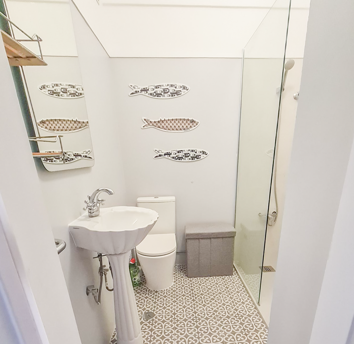 Small bathroom with a pedestal sink, a toilet, a gray storage box, and a glass shower door. Decor includes fish-shaped wall art and a mirror with fish patterns, with a patterned tile floor.