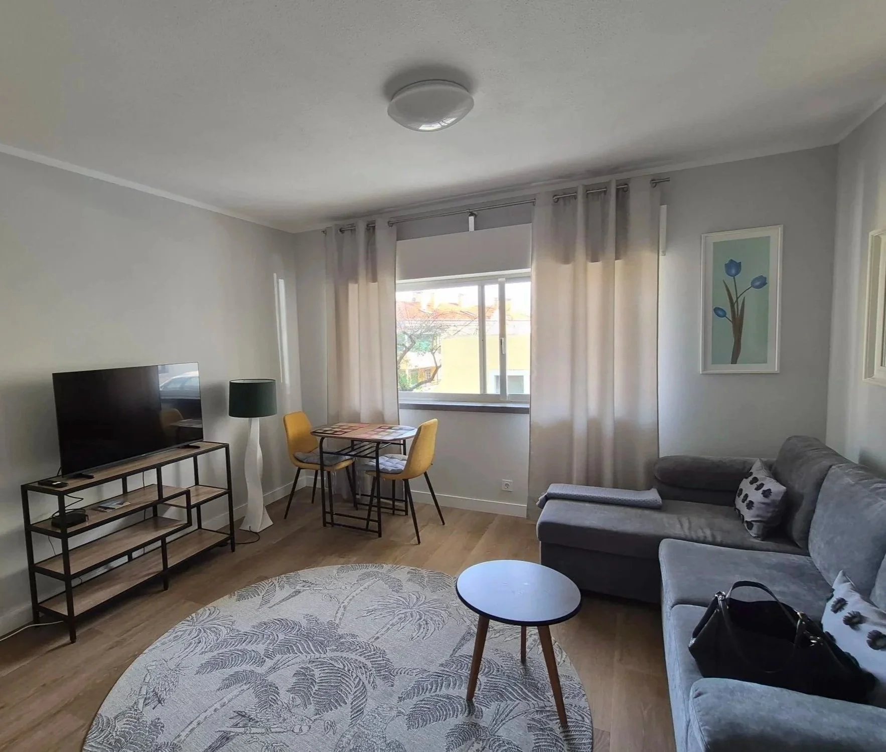 Living room with a gray sectional sofa, a TV on a black metal and wood stand, a potted plant, a window with white curtains, a patterned area rug, a round dining table with yellow chairs, and a framed picture on the wall.
