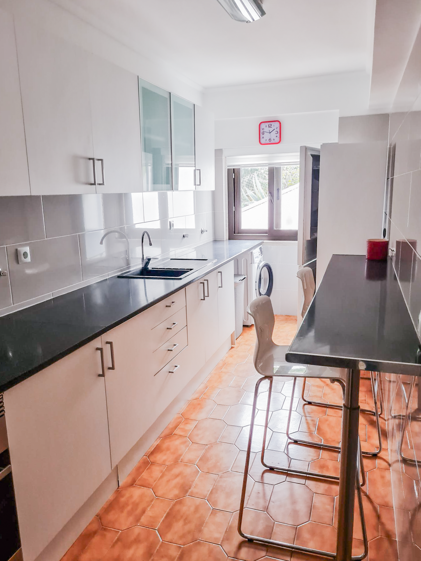 Bright kitchen with white cabinets, black countertop, orange tiled floor, washing machine, barstools at a black counter, and window with a red clock above.