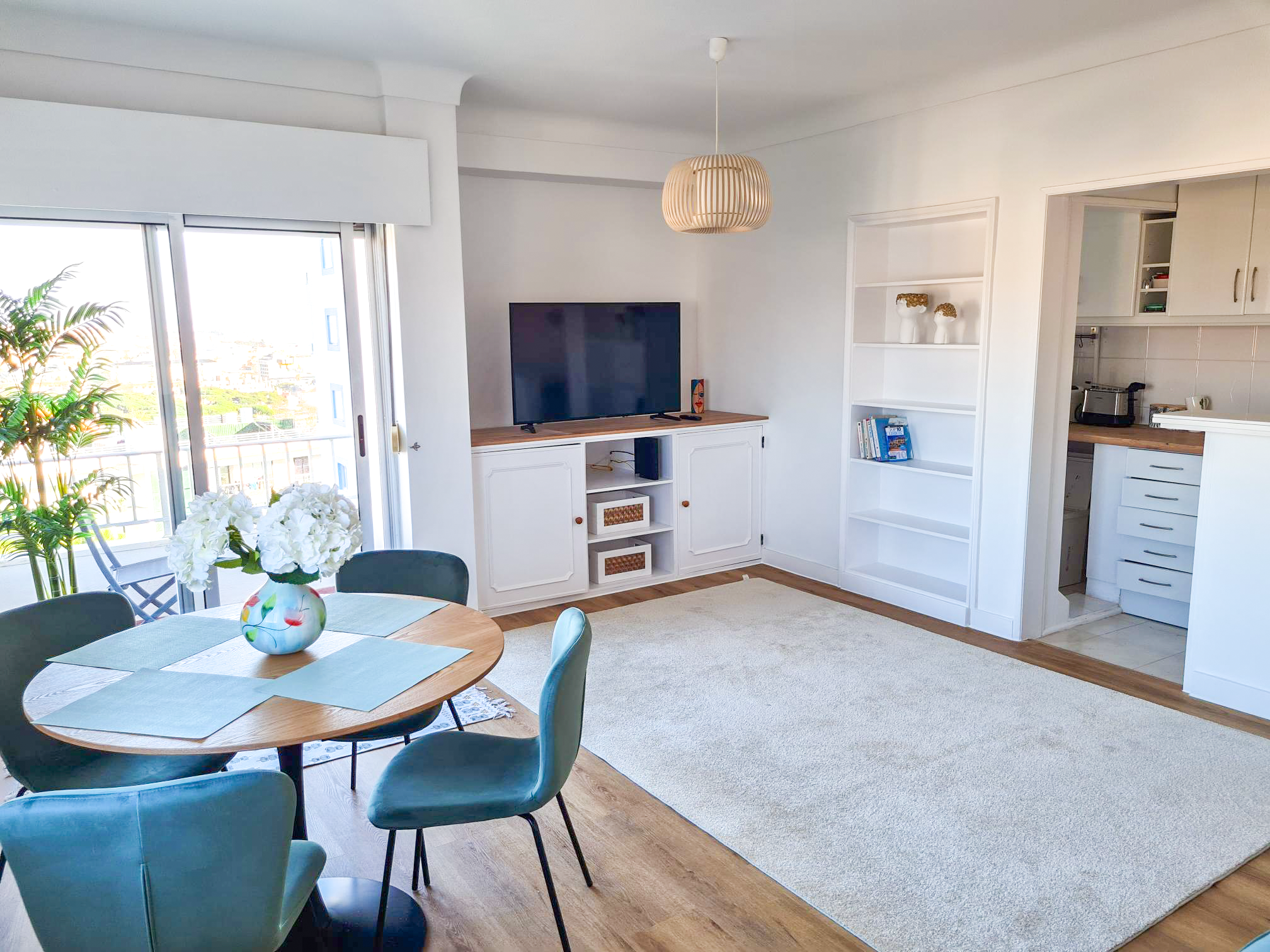 Living room with white walls, wooden floor, sliding glass door with balcony, round dining table with flowers, and a white TV stand with a TV
