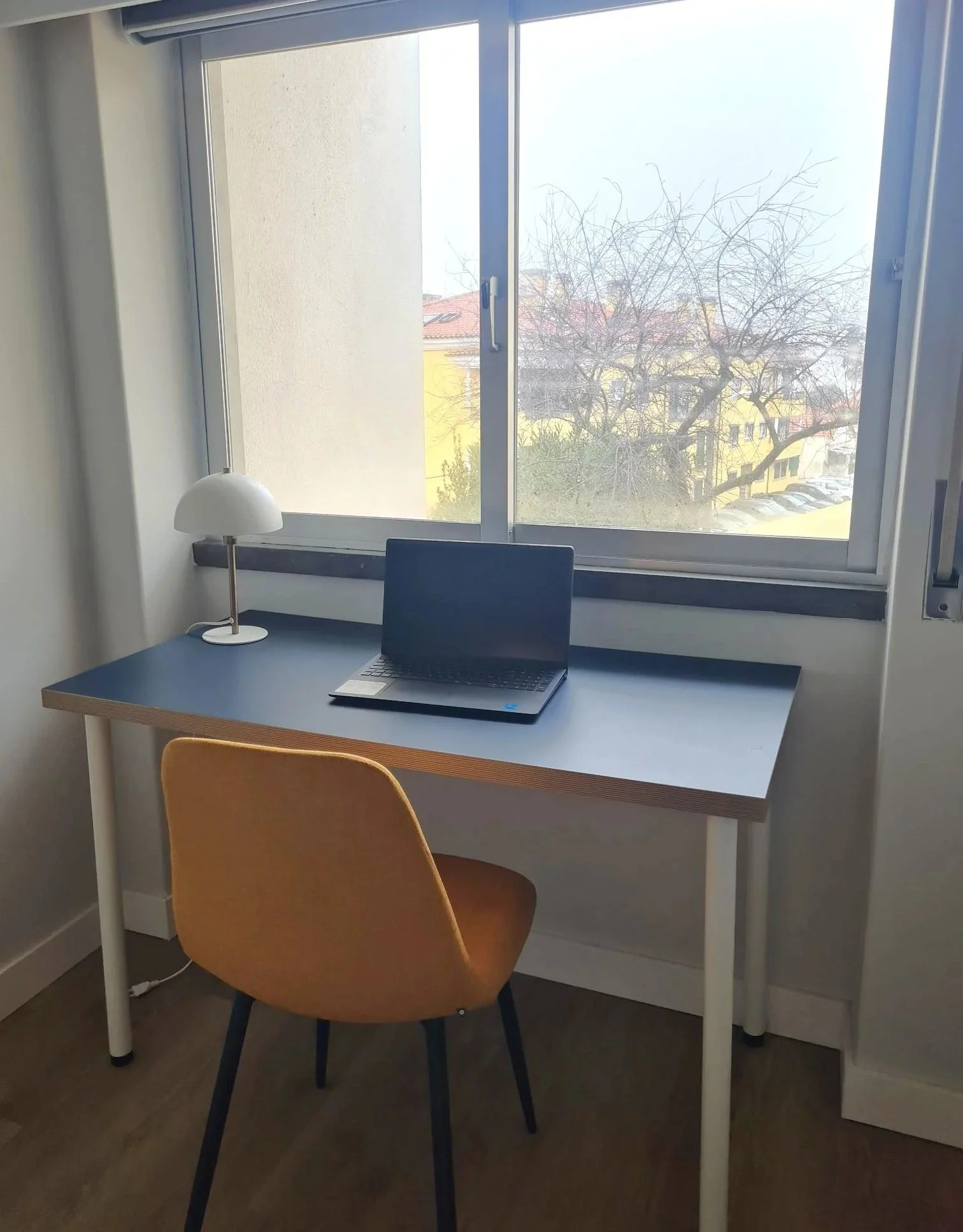 A simple workspace with a desk facing a window, a tan chair, a small white lamp, and a laptop on the desk. Outside the window, there are trees and buildings.